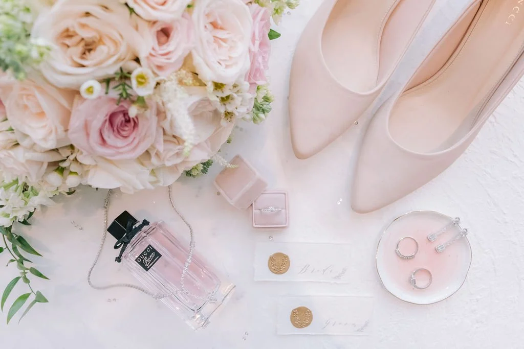 A flat lay of wedding or special occasion items including a bouquet of pink and white roses, a pair of nude-colored high heels, a bottle of Gucci perfume, a pink jewelry box, a pair of earrings, a ring, and two gold coins on a white surface.