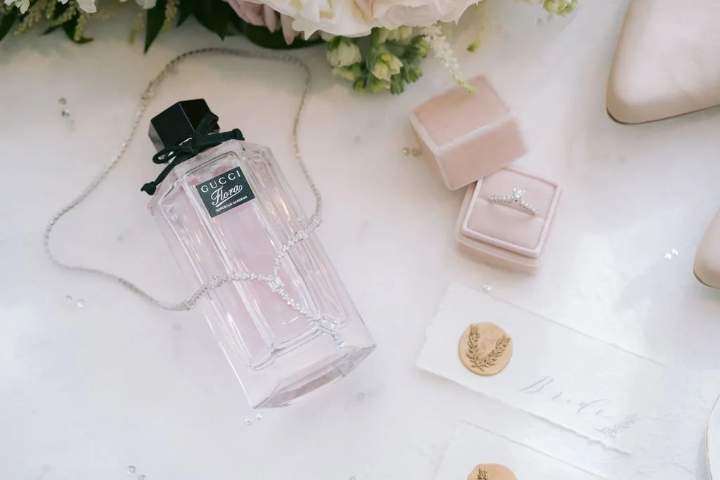 A bottle of Gucci Flora perfume, a silver necklace, a pink velvet jewelry box with a ring, and a small piece of paper with a gold wax seal on a white surface.