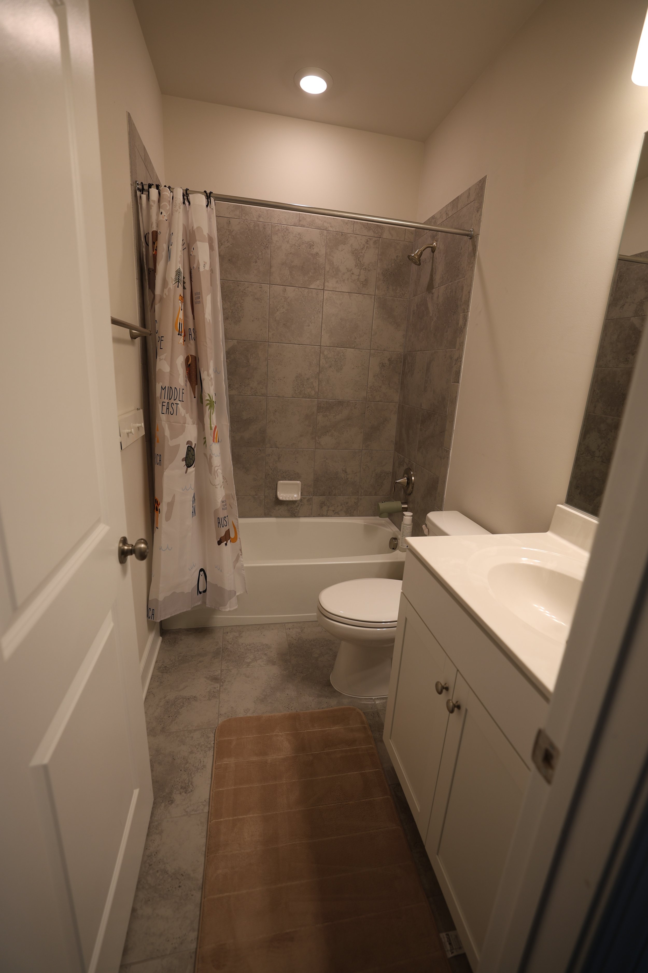 A small bathroom with a shower/tub combination, a toilet, a white vanity with a sink, a mirror, and a brown bath mat on the tiled floor. The shower has a beige tile surround and a shower curtain with animal illustrations.