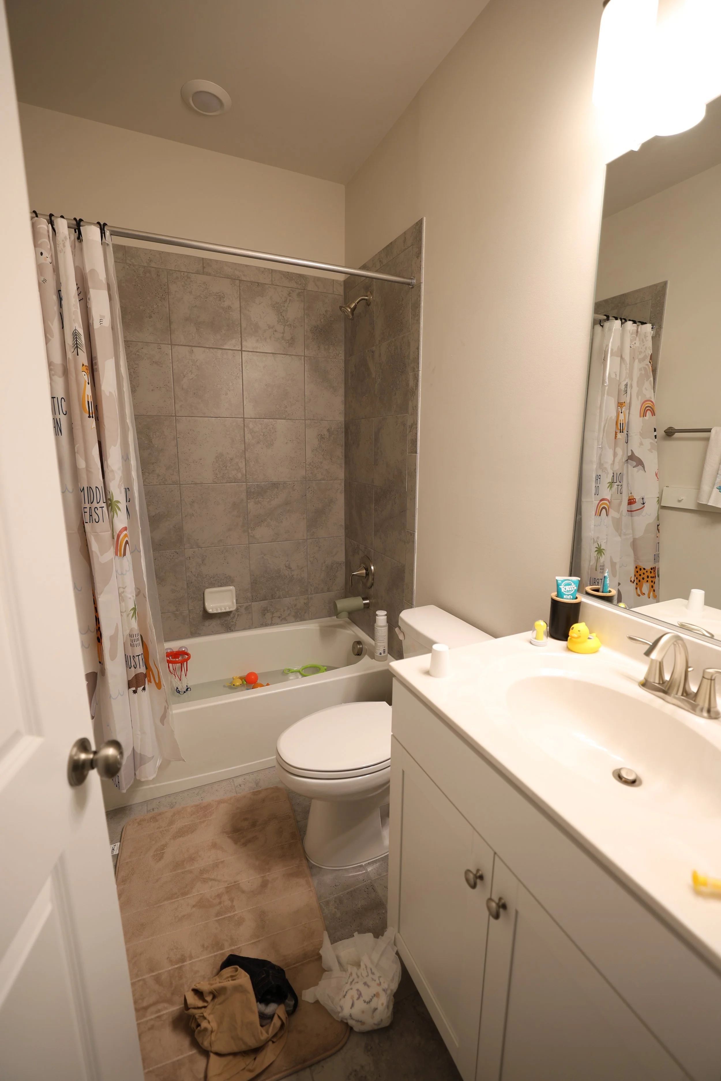 A small bathroom with a white vanity and mirror, a toilet, a bathtub with a tiled shower wall, and a shower curtain with animal illustrations. The floor has a brown bath mat, and there are children's toys, toiletries, and a towel on a rack.
