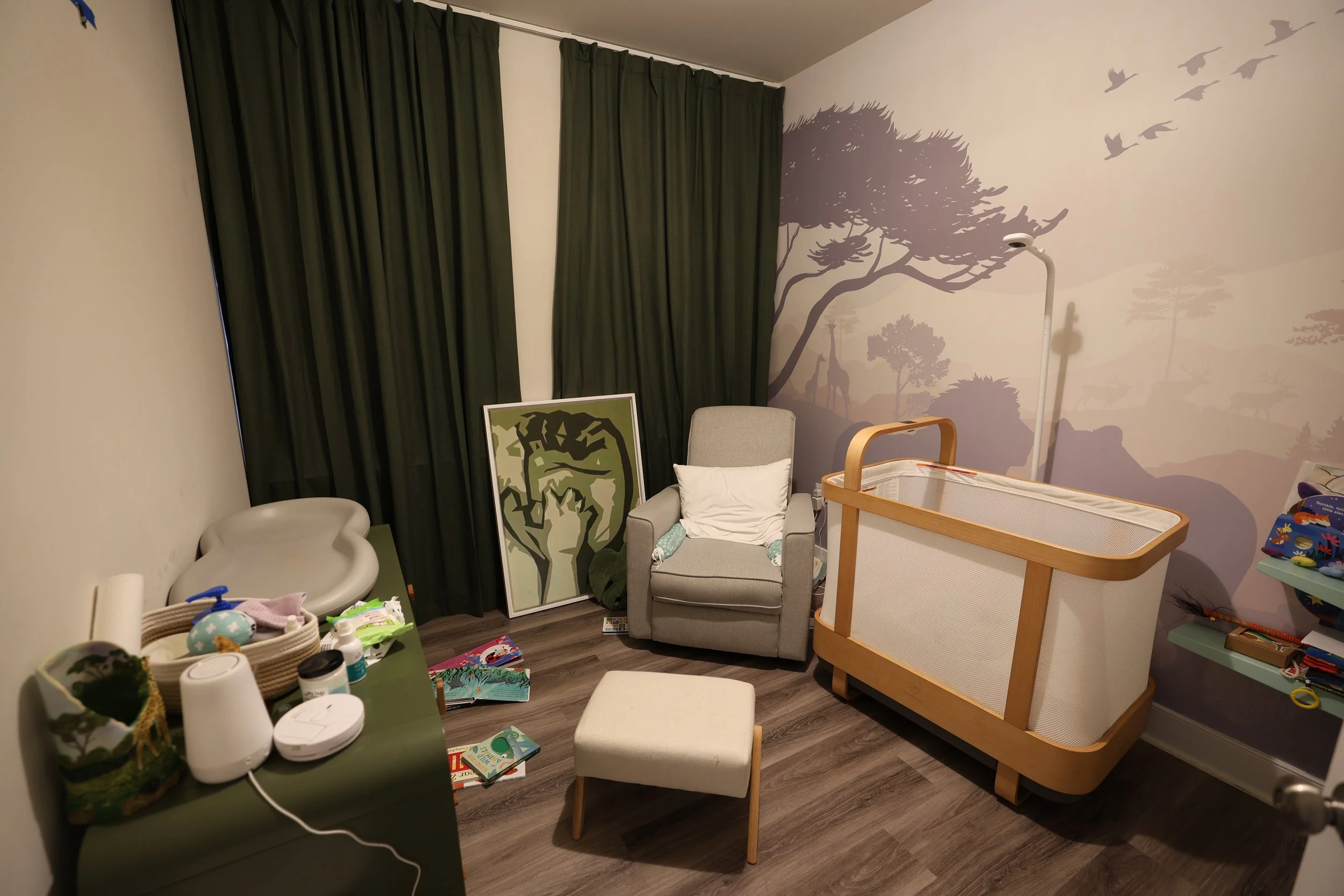 Nursery room with gray armchair, white pillow, large framed animal art leaning against wall, wooden crib, changing table with toys, wall mural of an African landscape with trees and giraffes, dark green curtains, small white ottoman, scattered childr