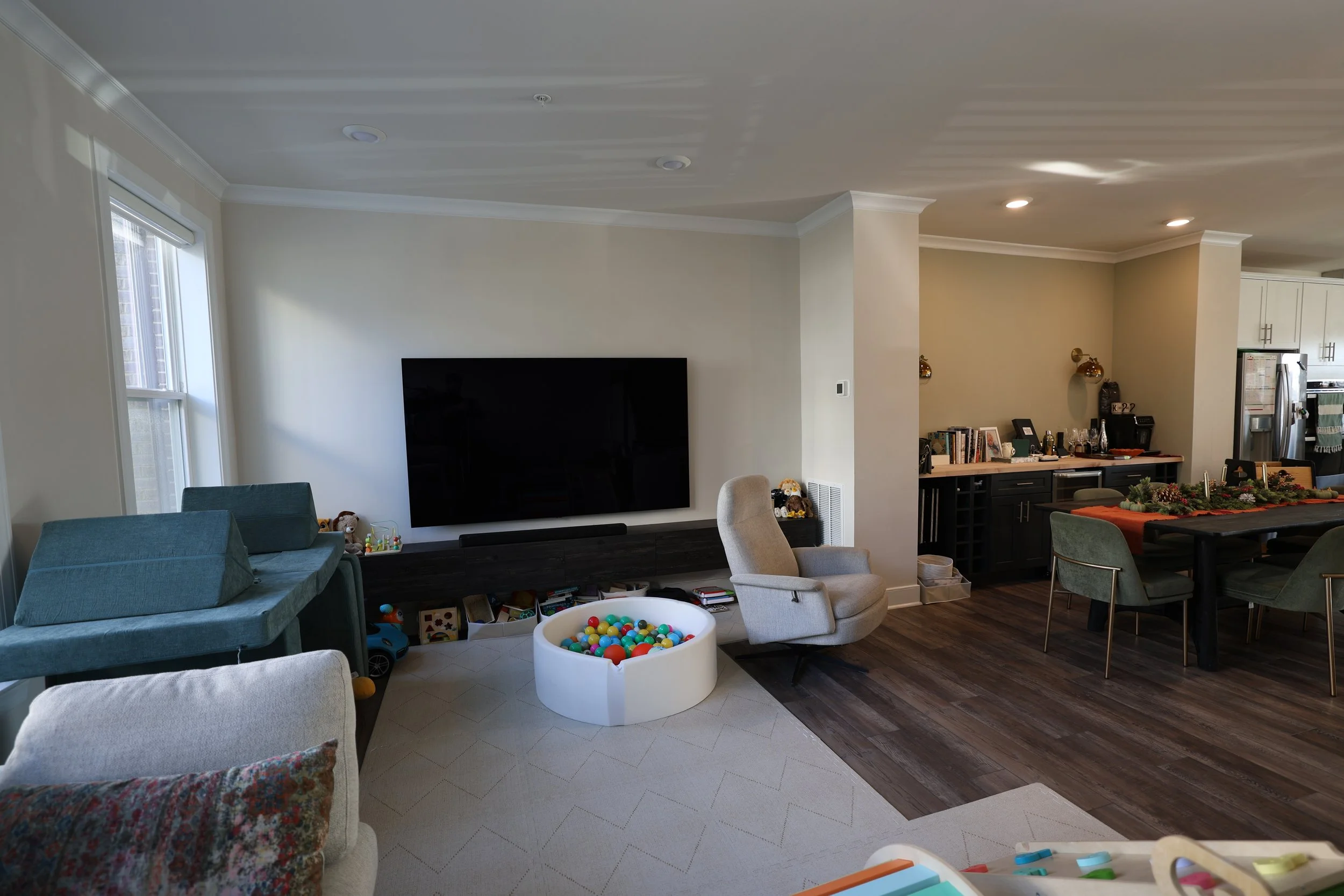 Living room with a large flat-screen TV on the wall, a white armchair, a blue sofa, a toy bin filled with colorful balls, and some children's toys on the floor. A dining table with holiday decor and green chairs is in the background, with kitchen app