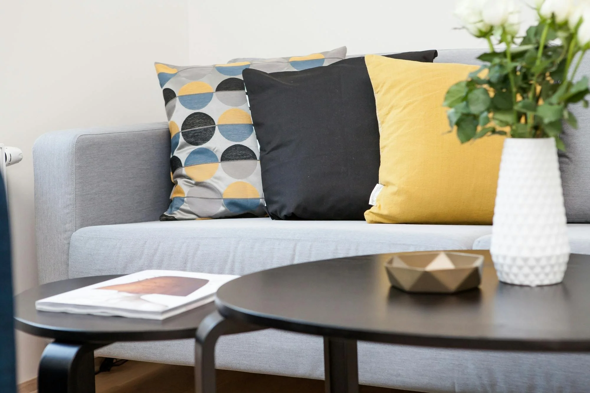 Living room sofa with three decorative pillows, one with a circular pattern in yellow, black, gray, and blue, another solid black, and the third solid yellow. A black coffee table with a magazine and decorative bowl is in front, and a white textured 