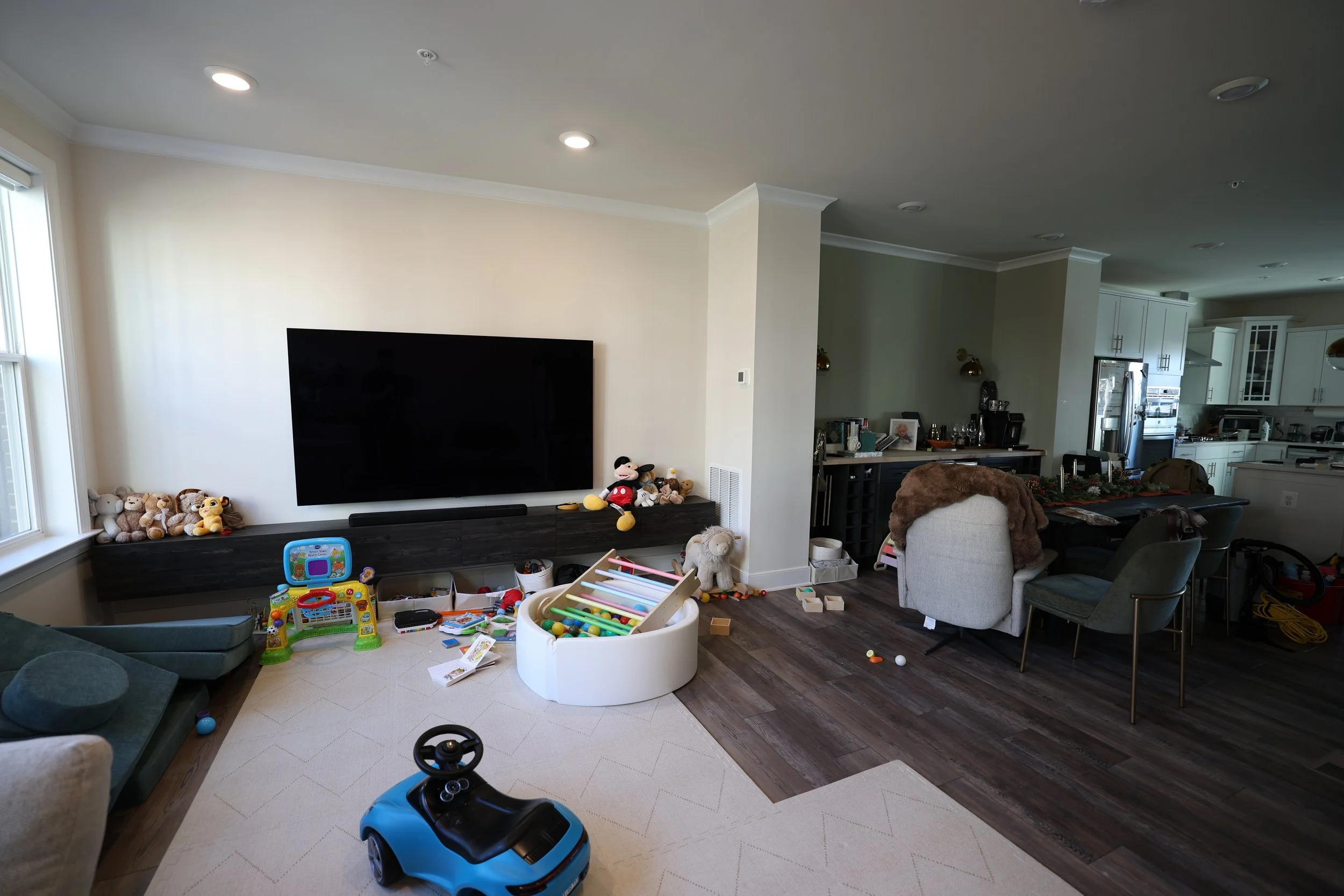 Living room with toys scattered on the floor, a large flat-screen TV on the wall, and a small child’s car on the carpet. Toys include stuffed animals, a colorful activity table, and various scattered balls and blocks. The room is connected to a kitch
