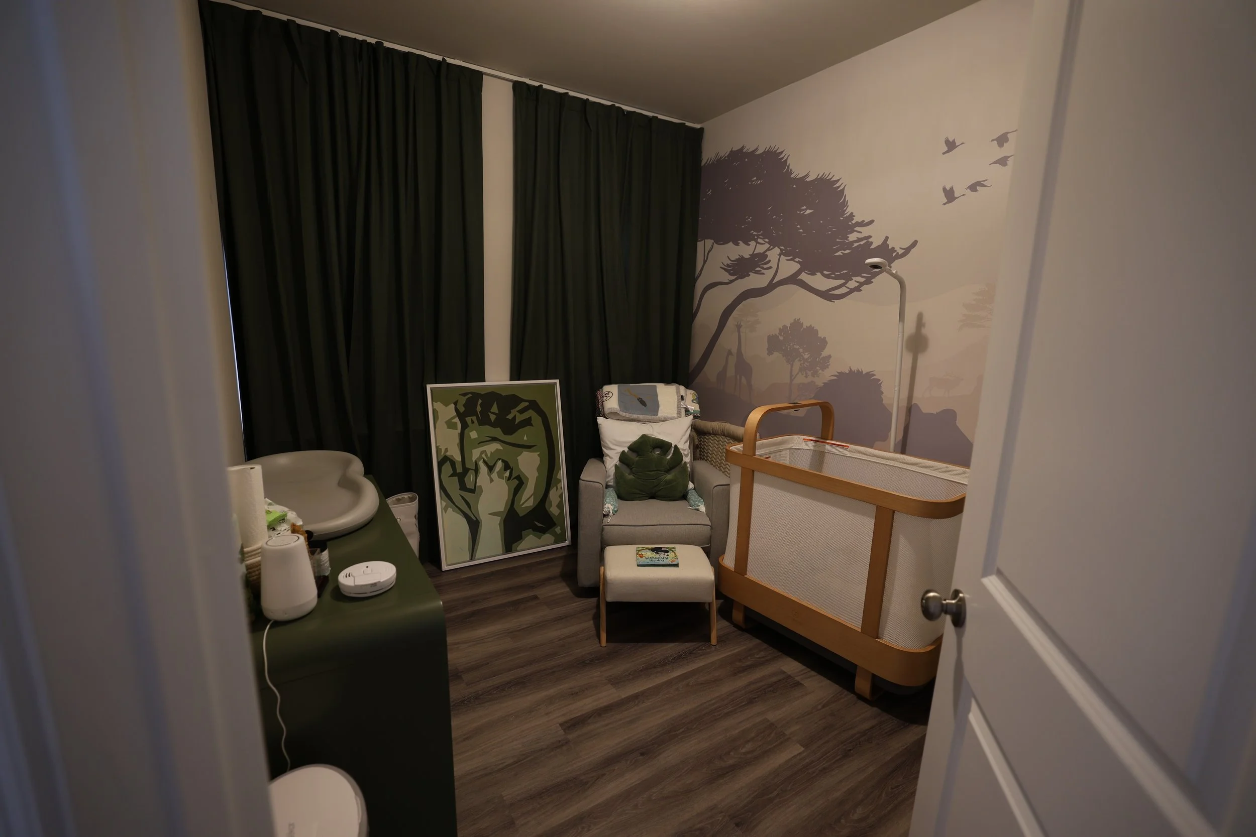 Nursery room with a mural of trees and birds on the wall, a chair with pillows, a changing table, and a framed artwork leaning against the wall. Contains dark green curtains and wood flooring.