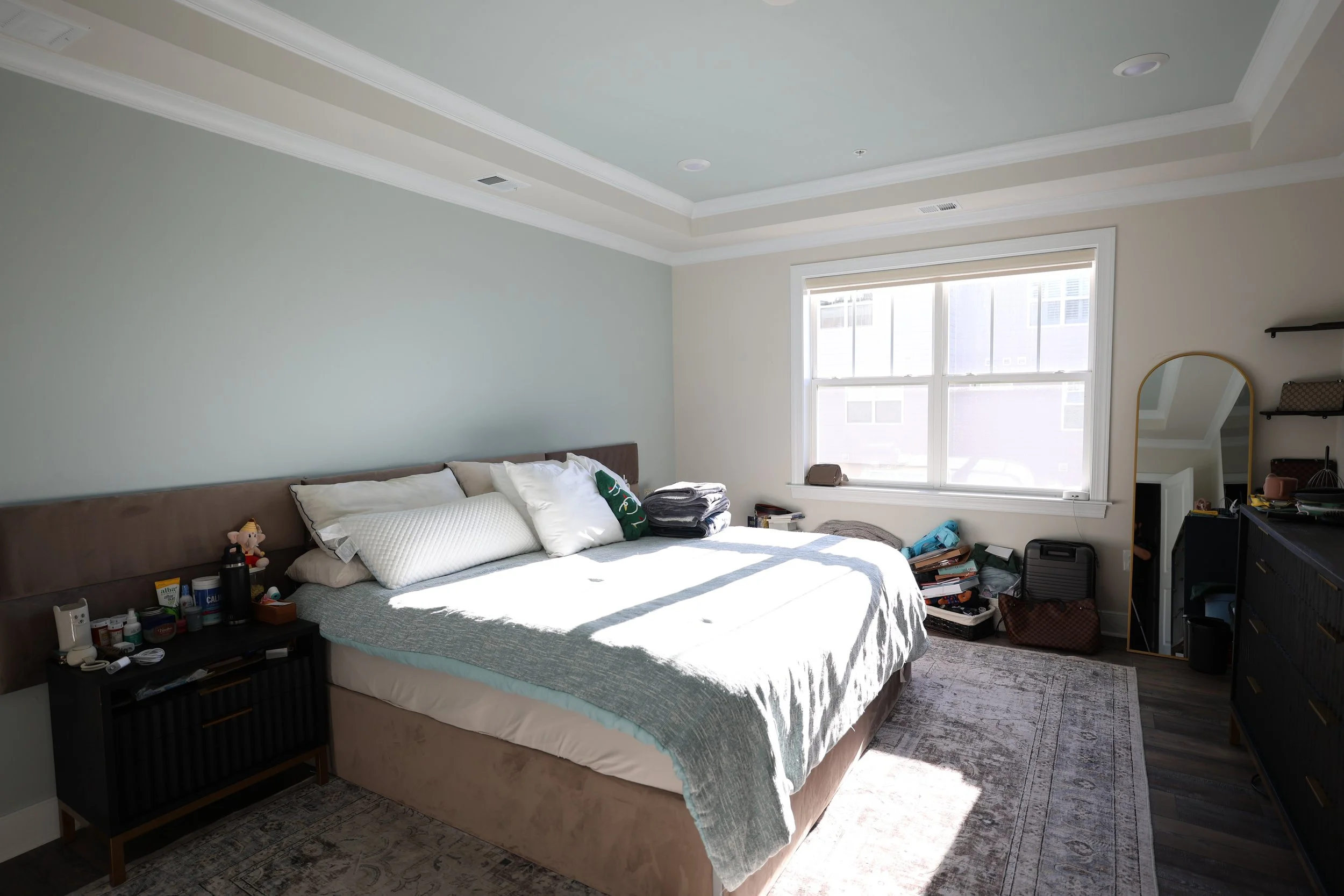 A bedroom with a large bed, nightstand, window, and dresser. The bed has white pillows and a blanket, with some items on it. The room is lit by natural light from the window, and there are various items and clutter near the window and on the dresser.
