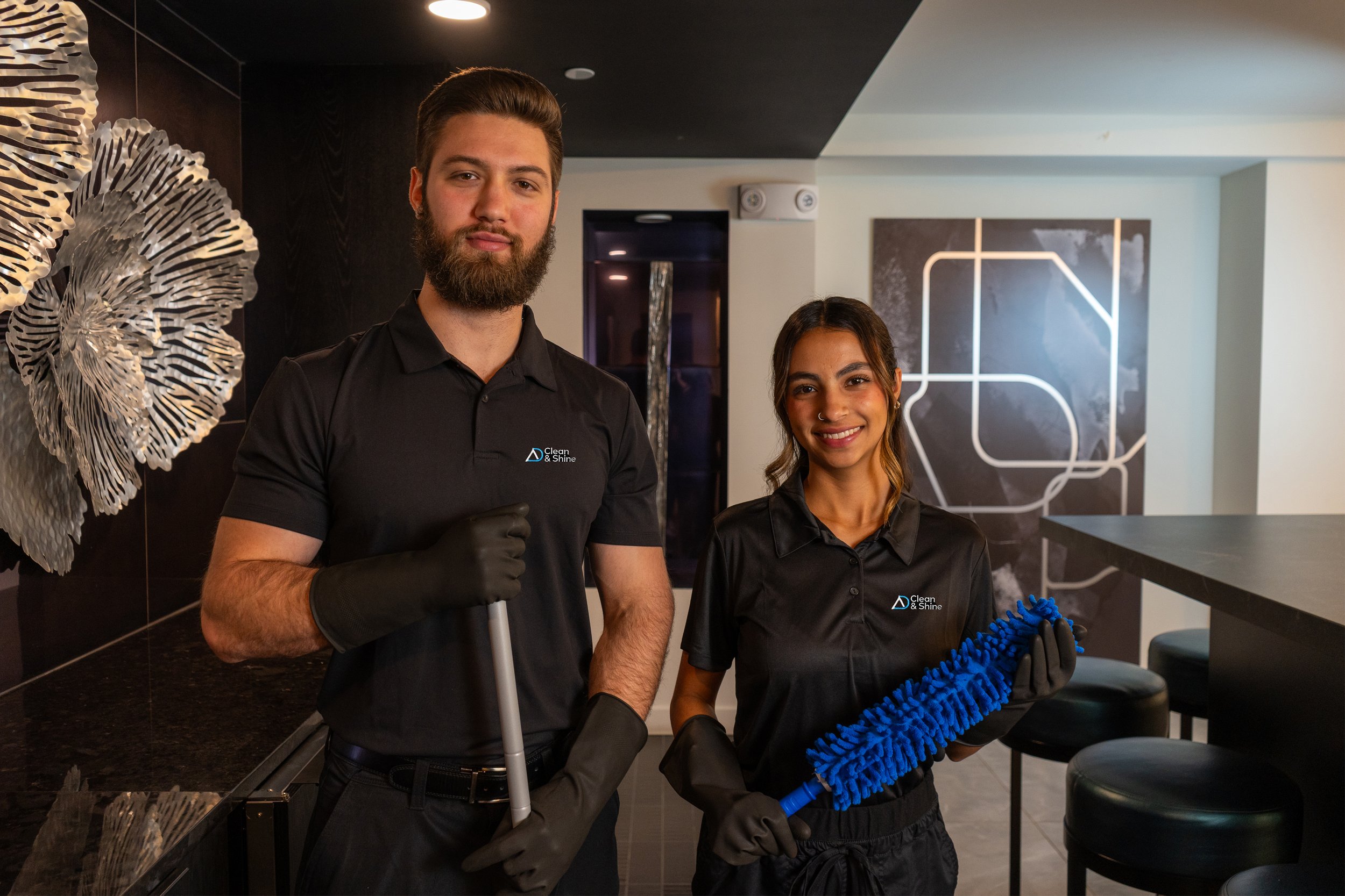 Two cleaning staff members in black uniforms standing in a modern, stylish interior, holding cleaning tools, smiling at the camera.