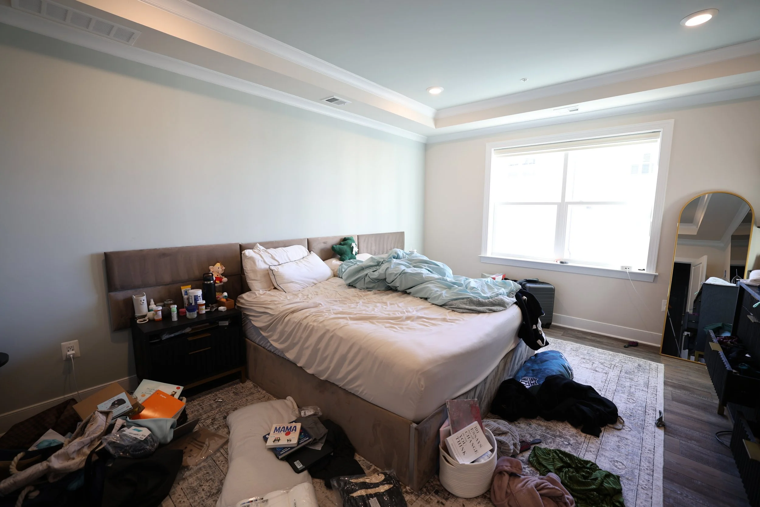 Disheveled bedroom with an unmade bed, cluttered floor, and a large window in the background.