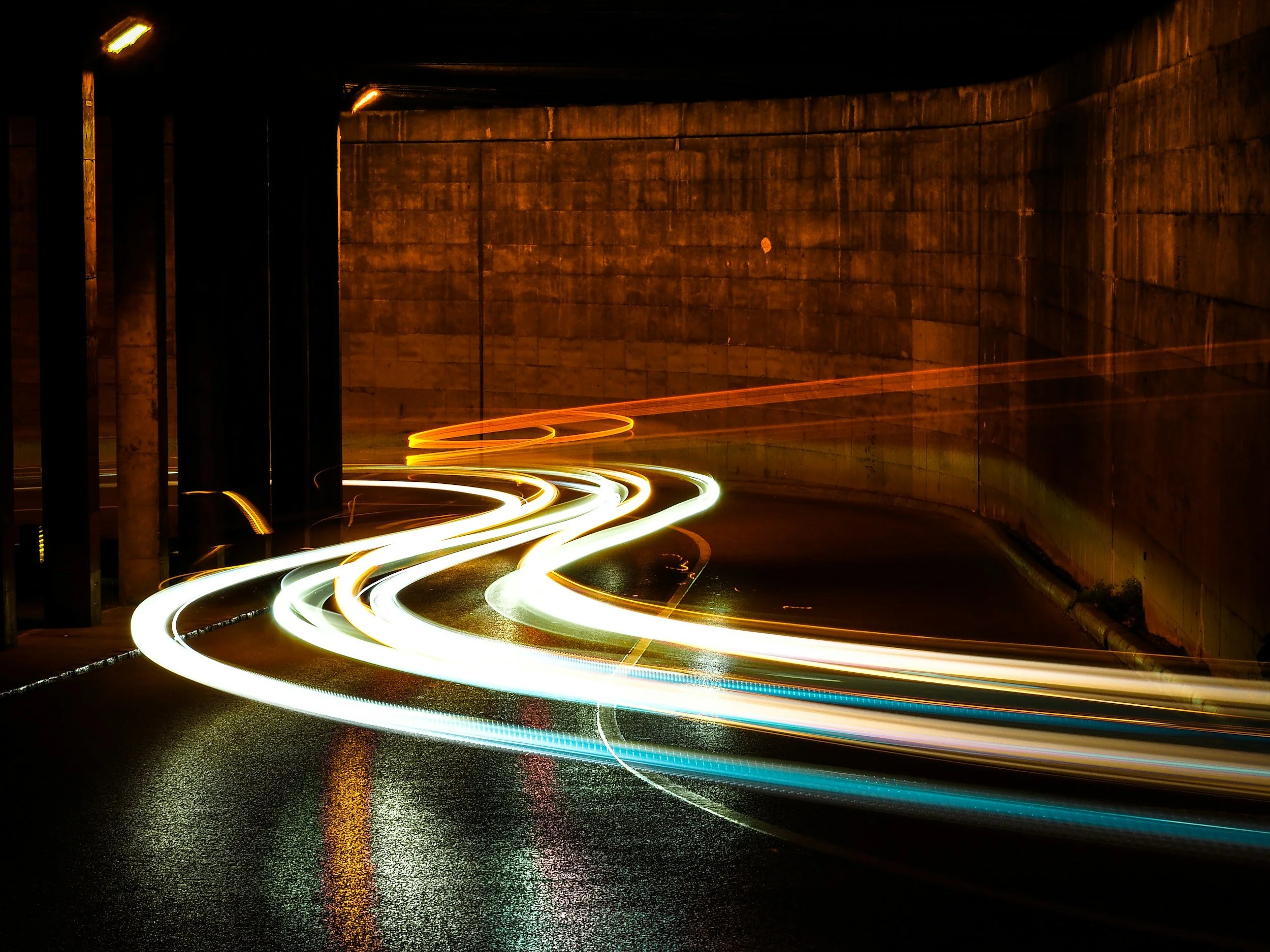 Light trails created by moving vehicle headlights on a curved, wet road in a tunnel at night.