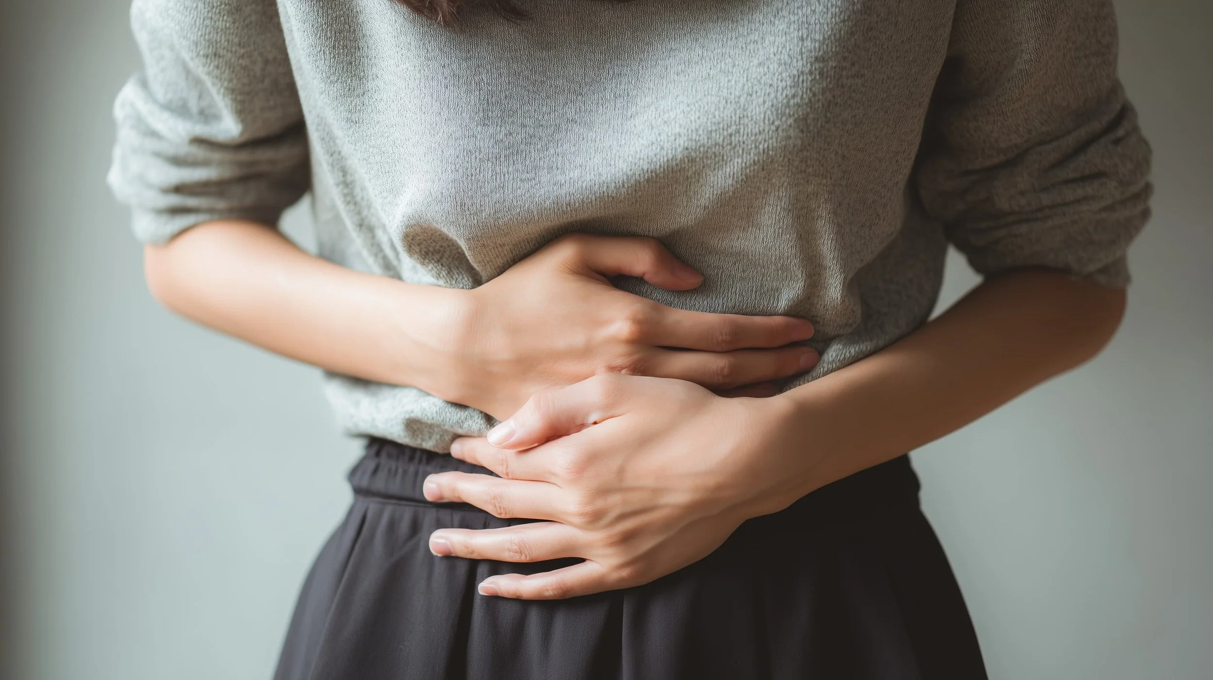 Woman holding her lower abdomen with both hands, representing abdominal pain or stomach discomfort, soft natural lighting and neutral background.