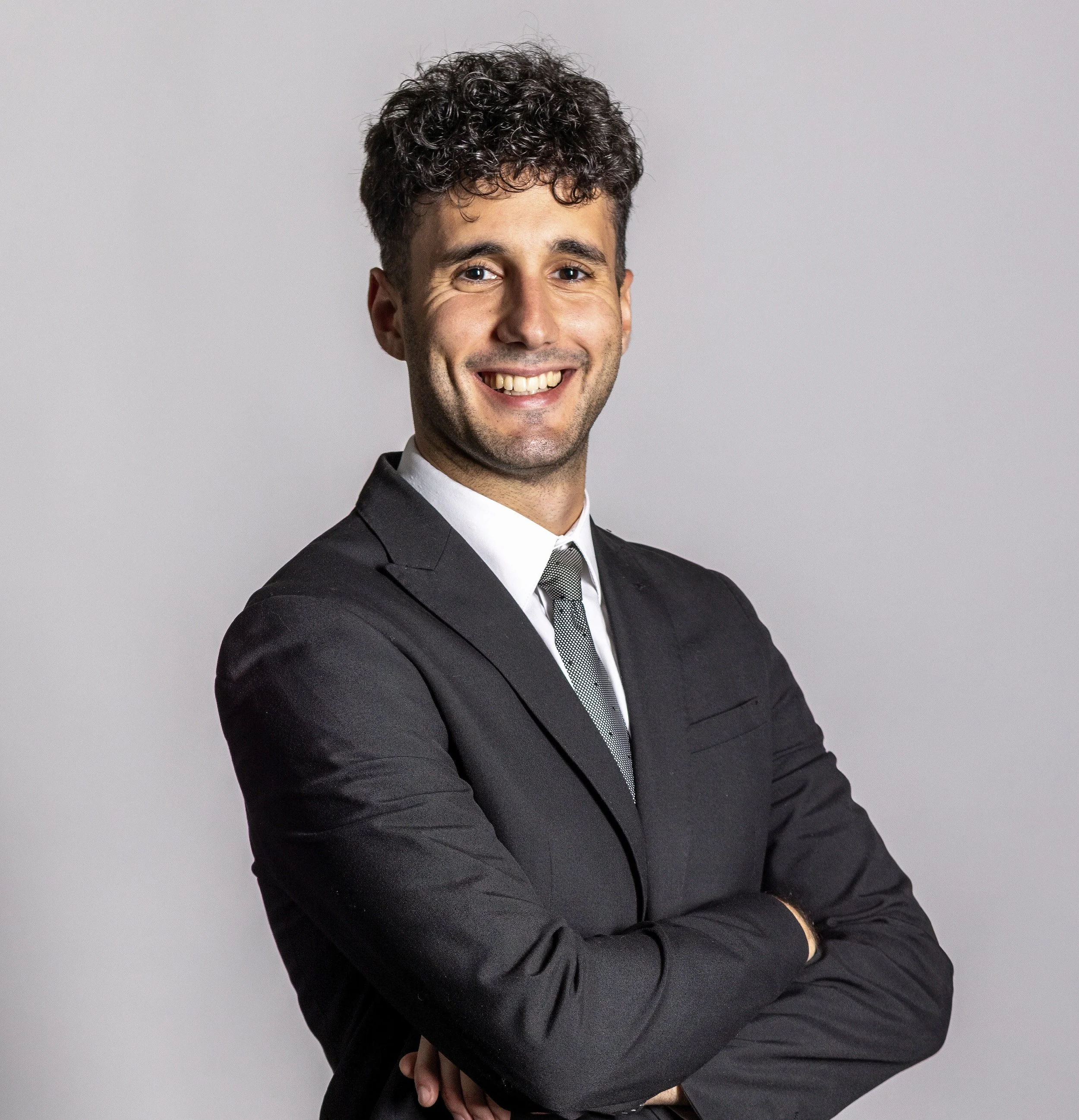 Uomo sorridente con capelli ricci, indossa un abito elegante nero e una cravatta, sfondo grigio.