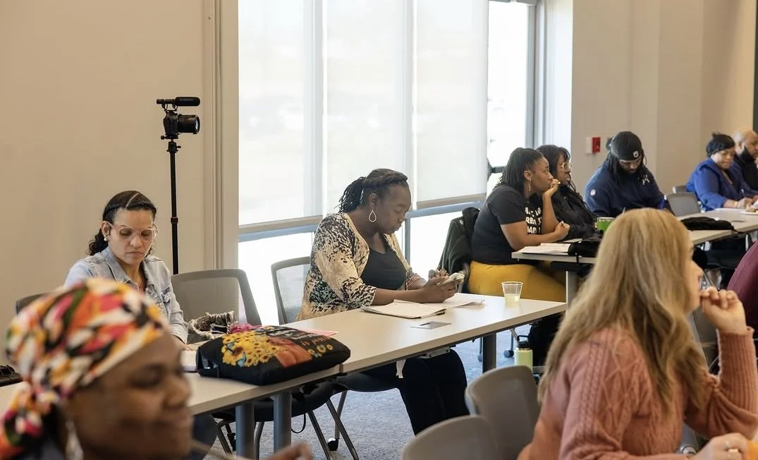A diverse group of women attending a meeting or workshop in a well-lit conference room with large windows.