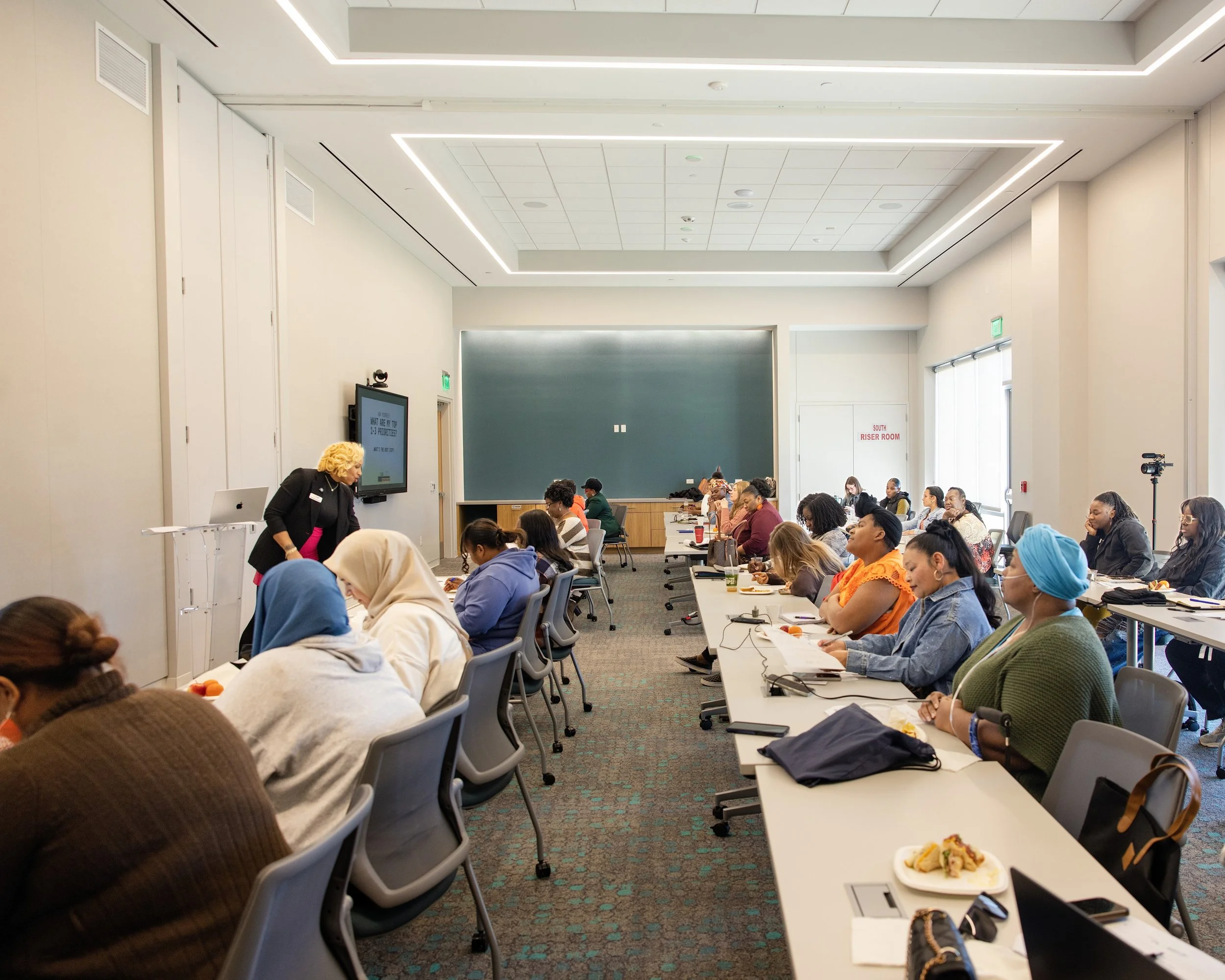 A classroom with diverse students seated at long white tables, attentively listening to an instructor at the front. The instructor stands near a digital display and a chalkboard, with a laptop on a stand. The room has a high ceiling with modern lighting, windows on the right, and a sign that reads 'Riser Room' on the door.