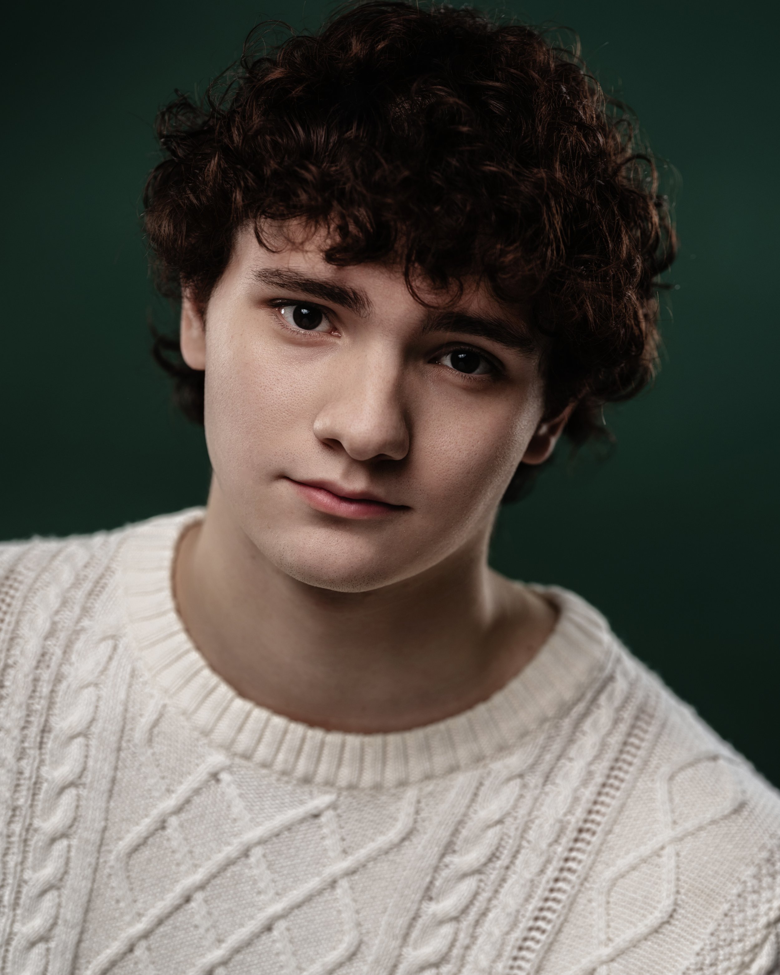 A young man with curly brown hair wearing a cream cable-knit sweater against a dark green background.