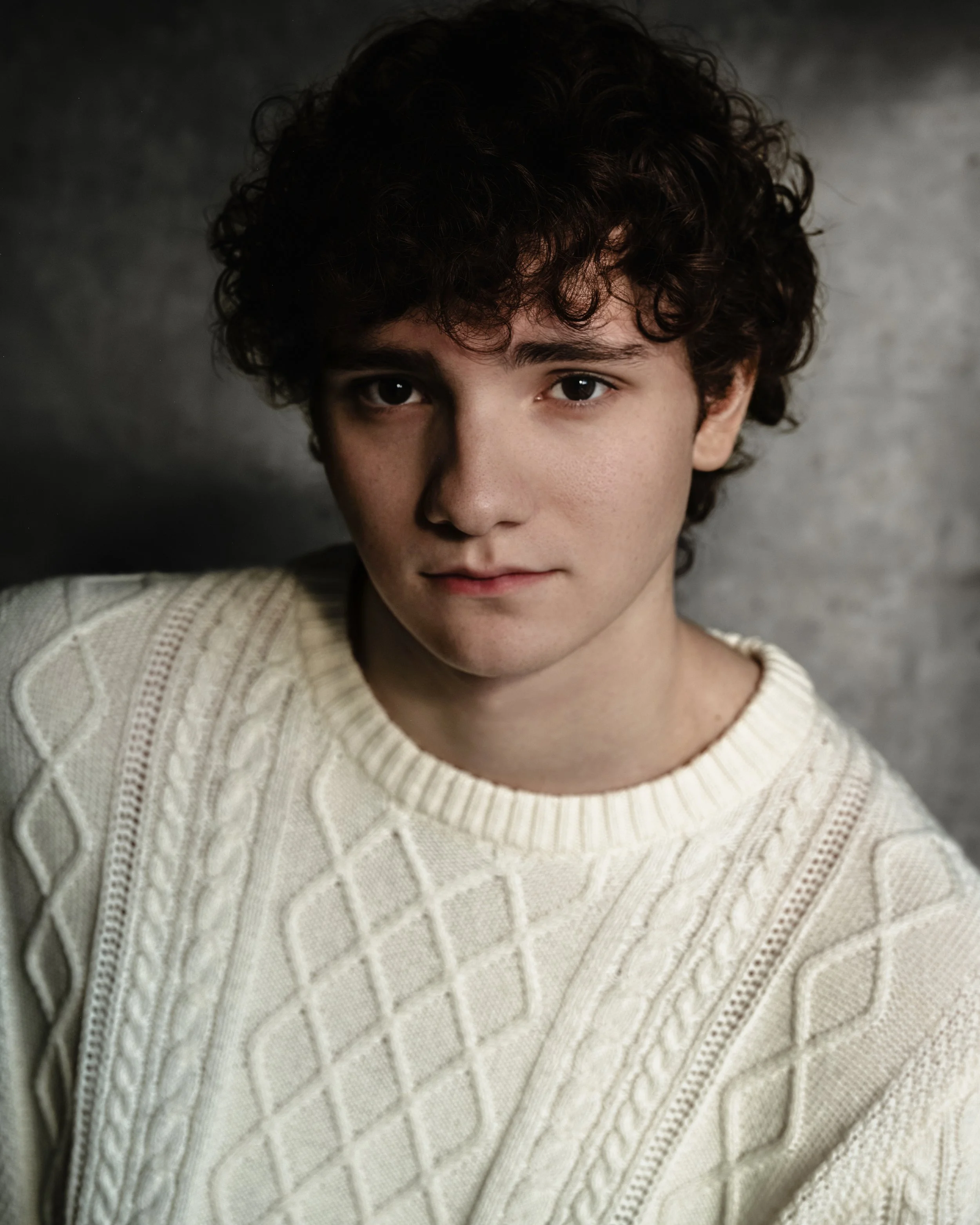A young man with curly dark hair wearing a cream-colored, textured knit sweater, looking directly at the camera with a neutral expression.