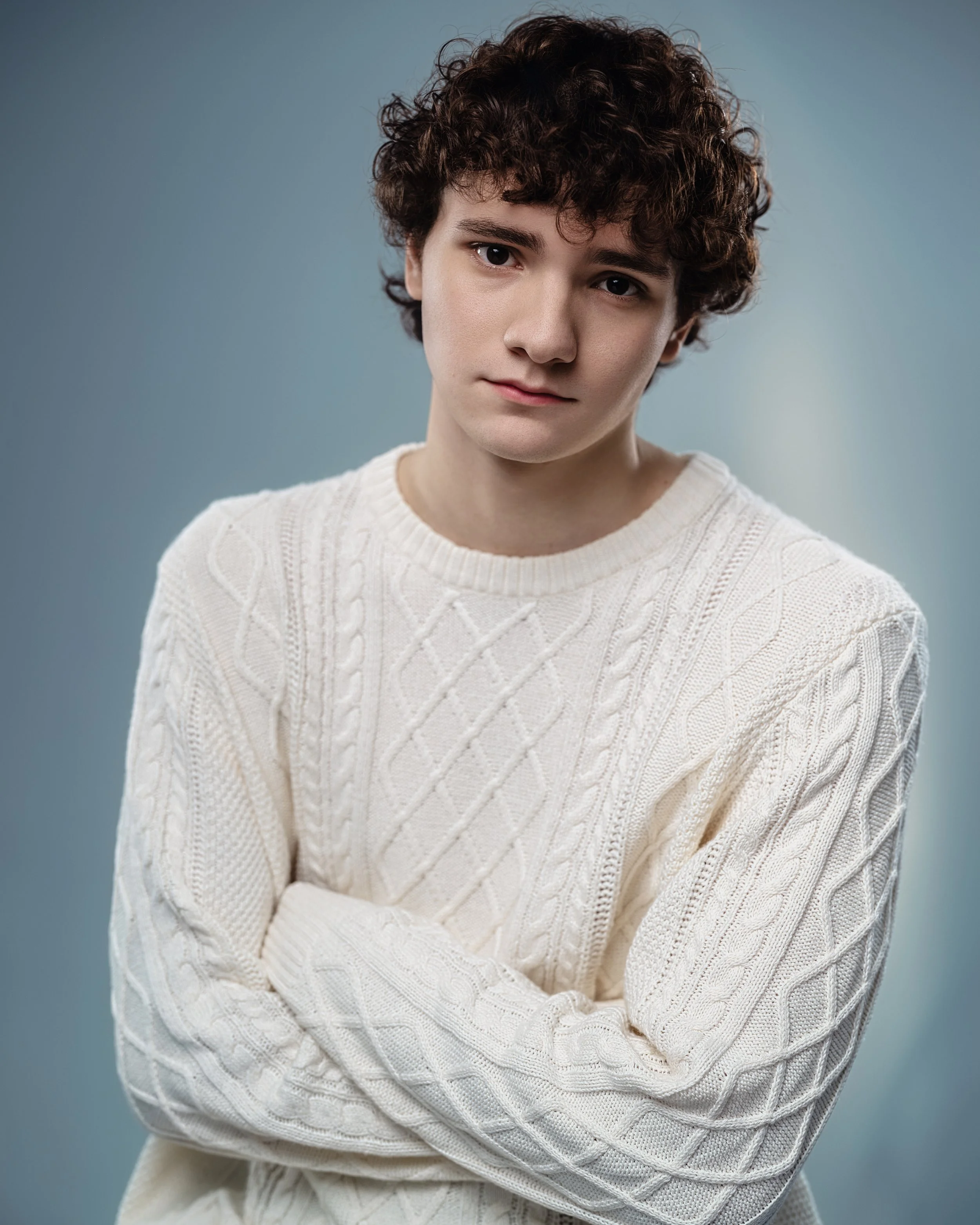 Young man with curly brown hair wearing a cream cable-knit sweater, arms crossed, looking at the camera against a neutral background.