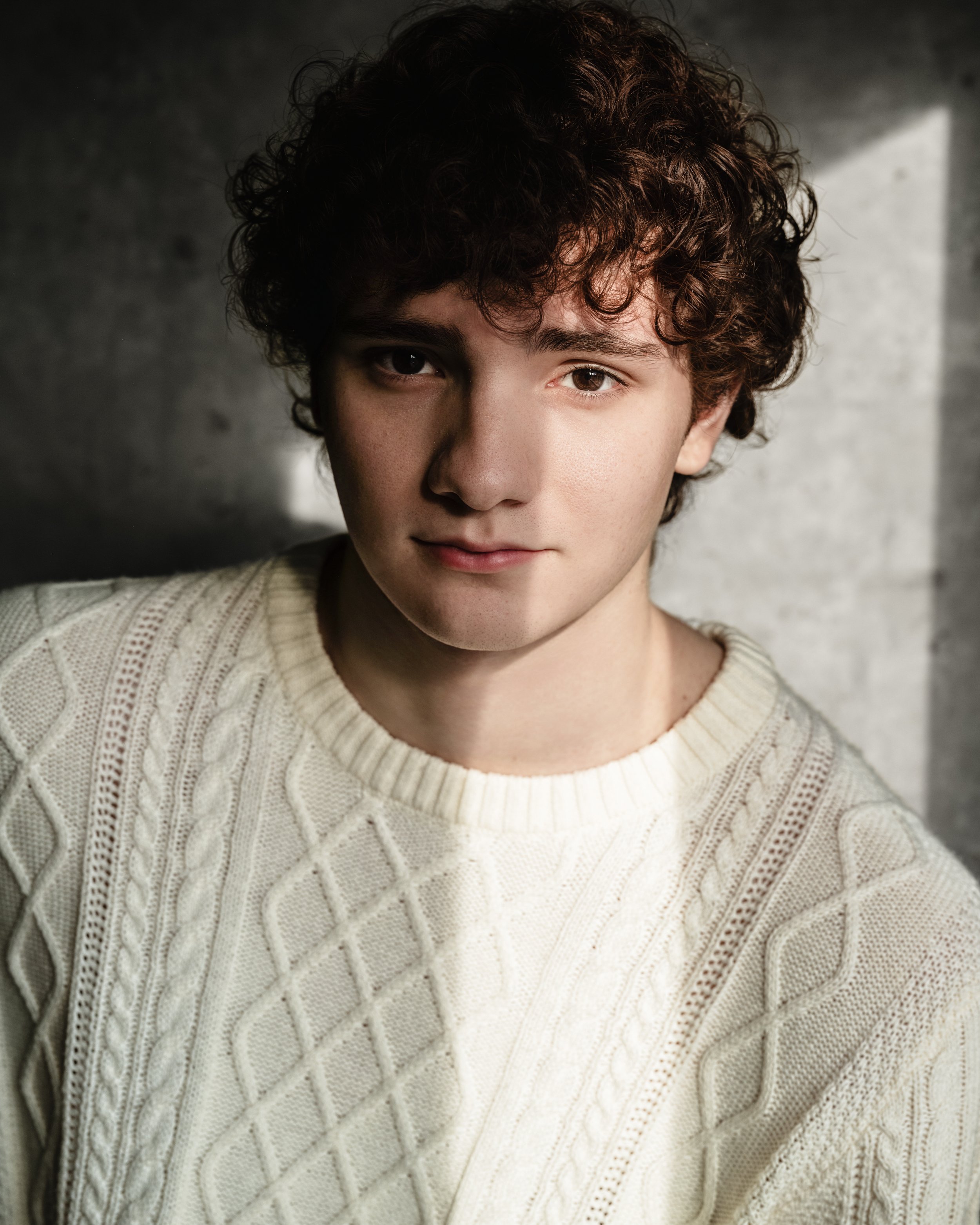 Portrait of a young man with curly brown hair, wearing a cream cable-knit sweater, standing against a gray wall with a soft light.
