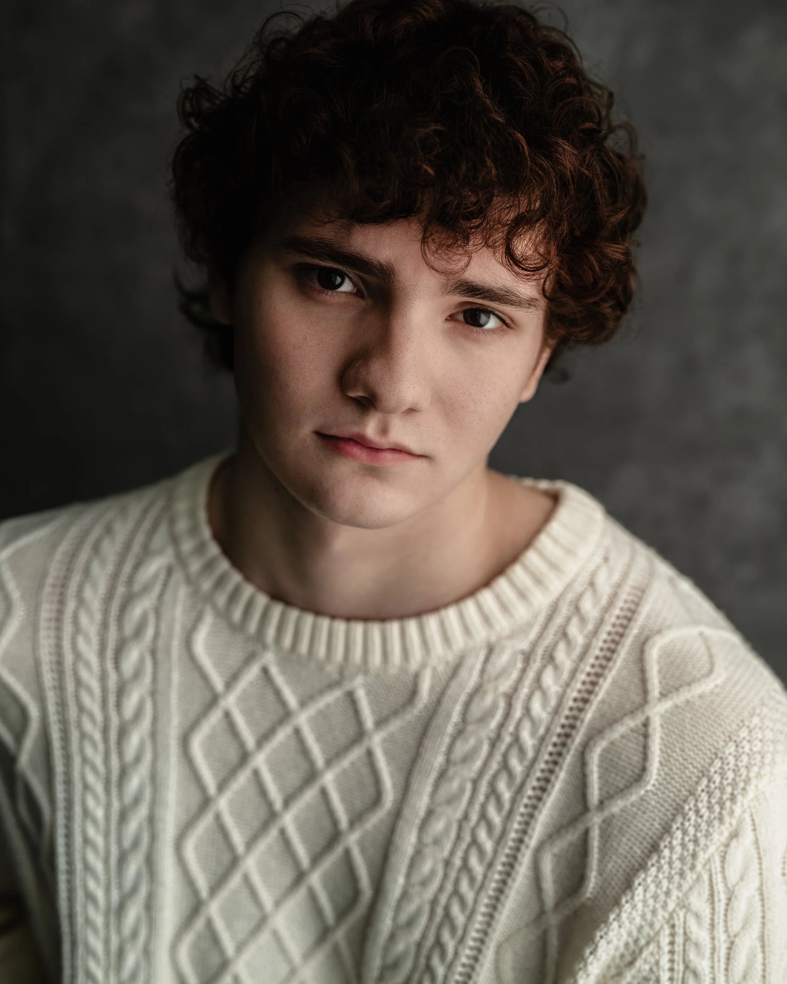 A young man with curly brown hair wearing a cream cable-knit sweater, looking directly at the camera against a dark gray background.