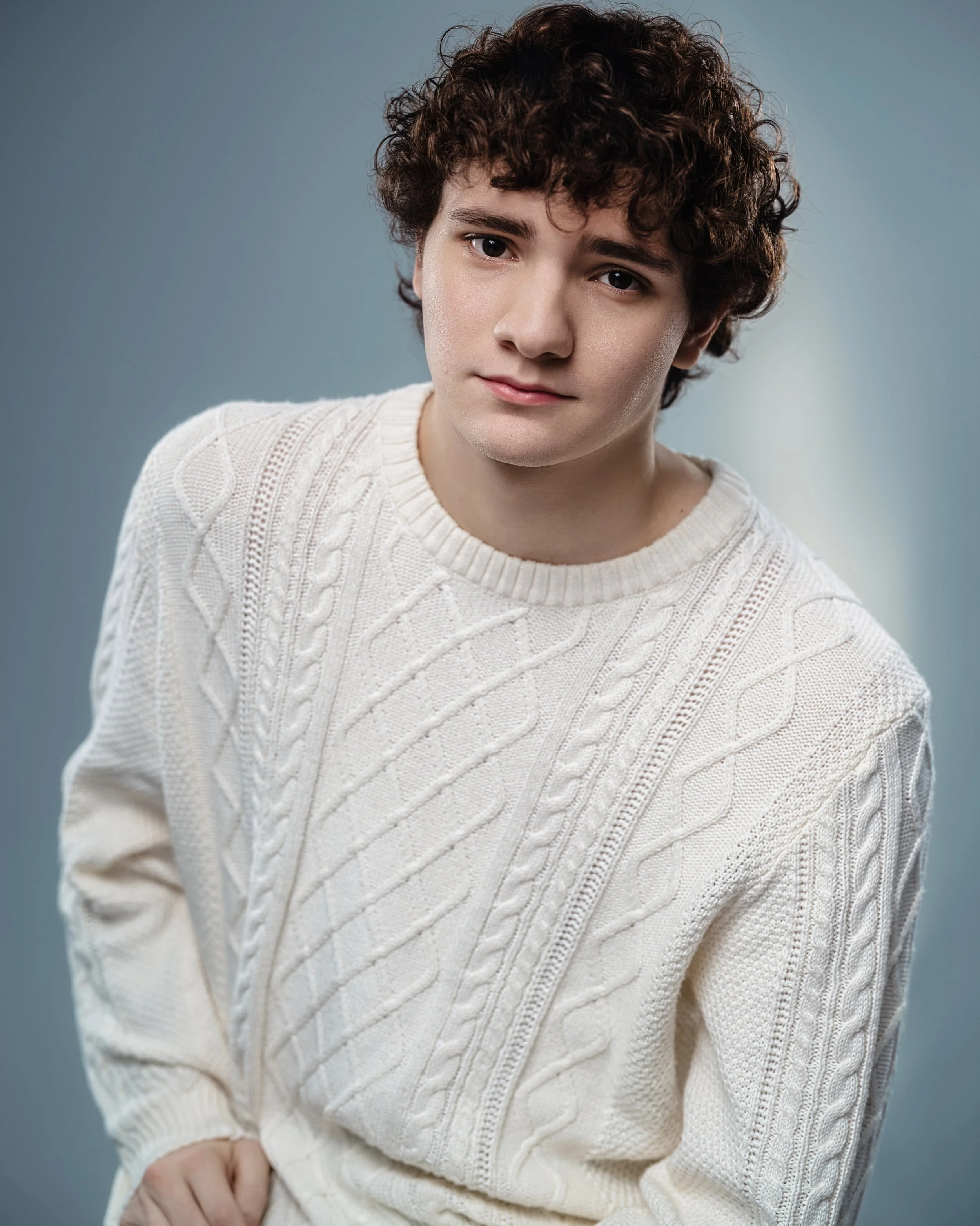 A young man with curly brown hair and fair skin, wearing a white cable-knit sweater, looking at the camera with a neutral expression against a plain gray background.