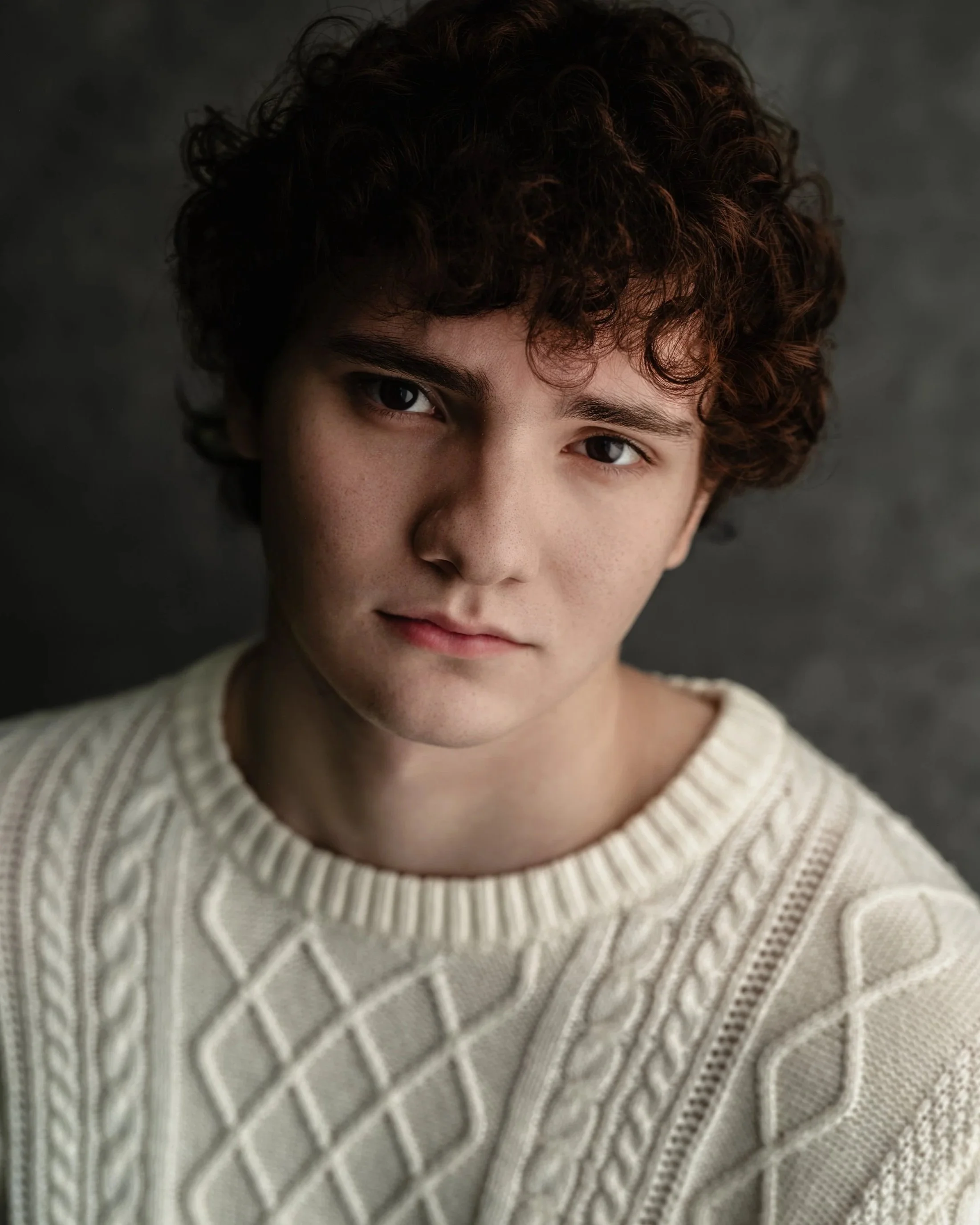 A young person with curly brown hair wearing a cream-colored knitted sweater, looking at the camera against a dark background.