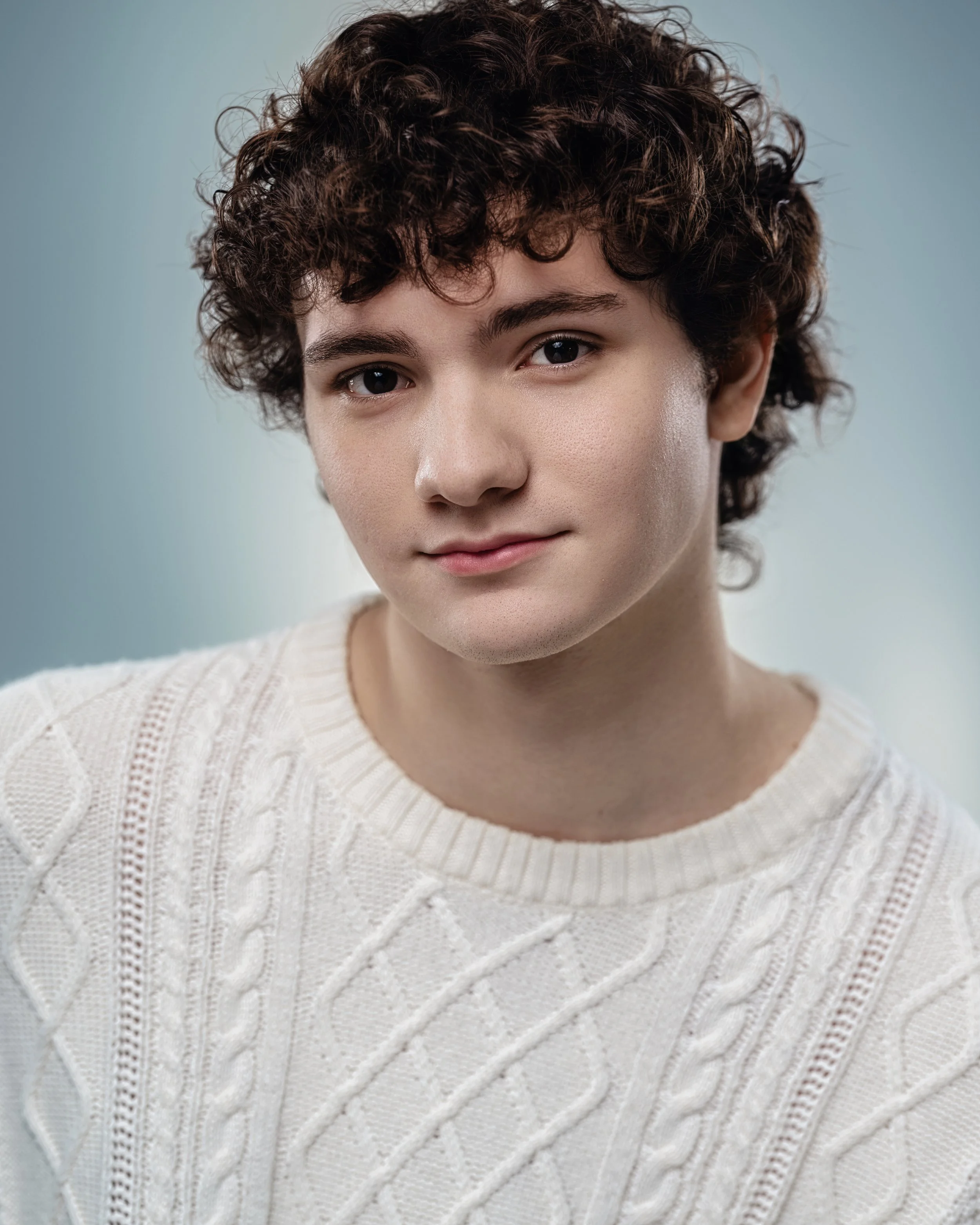 A young person with dark curly hair wearing a white knitted sweater, looking at the camera with a slight smile.