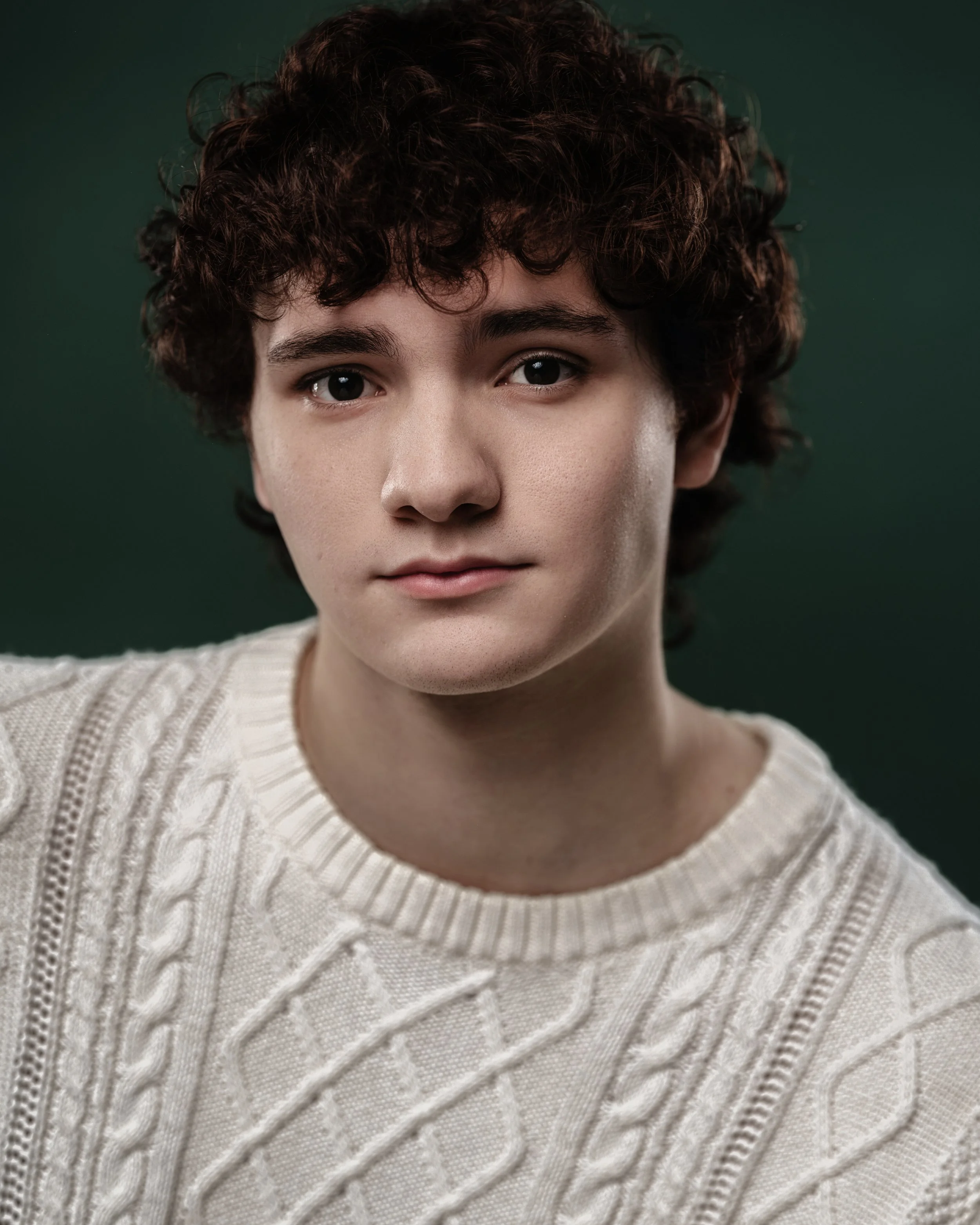 Close-up of a young person with curly dark hair, wearing a beige cable-knit sweater, looking into the camera with a neutral expression against a dark green background.