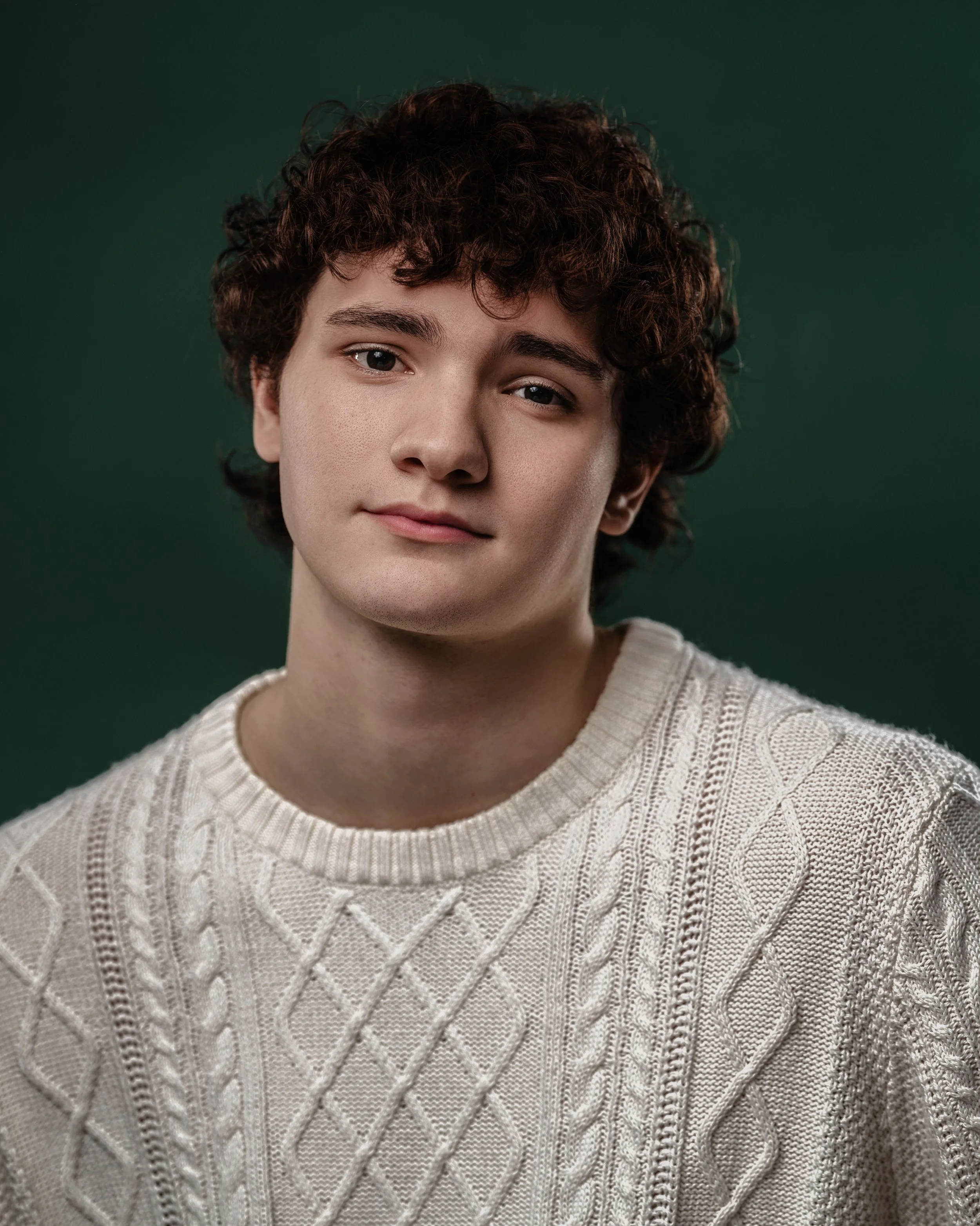 A young man with curly brown hair wearing a cream-colored cable knit sweater, standing against a dark green background.