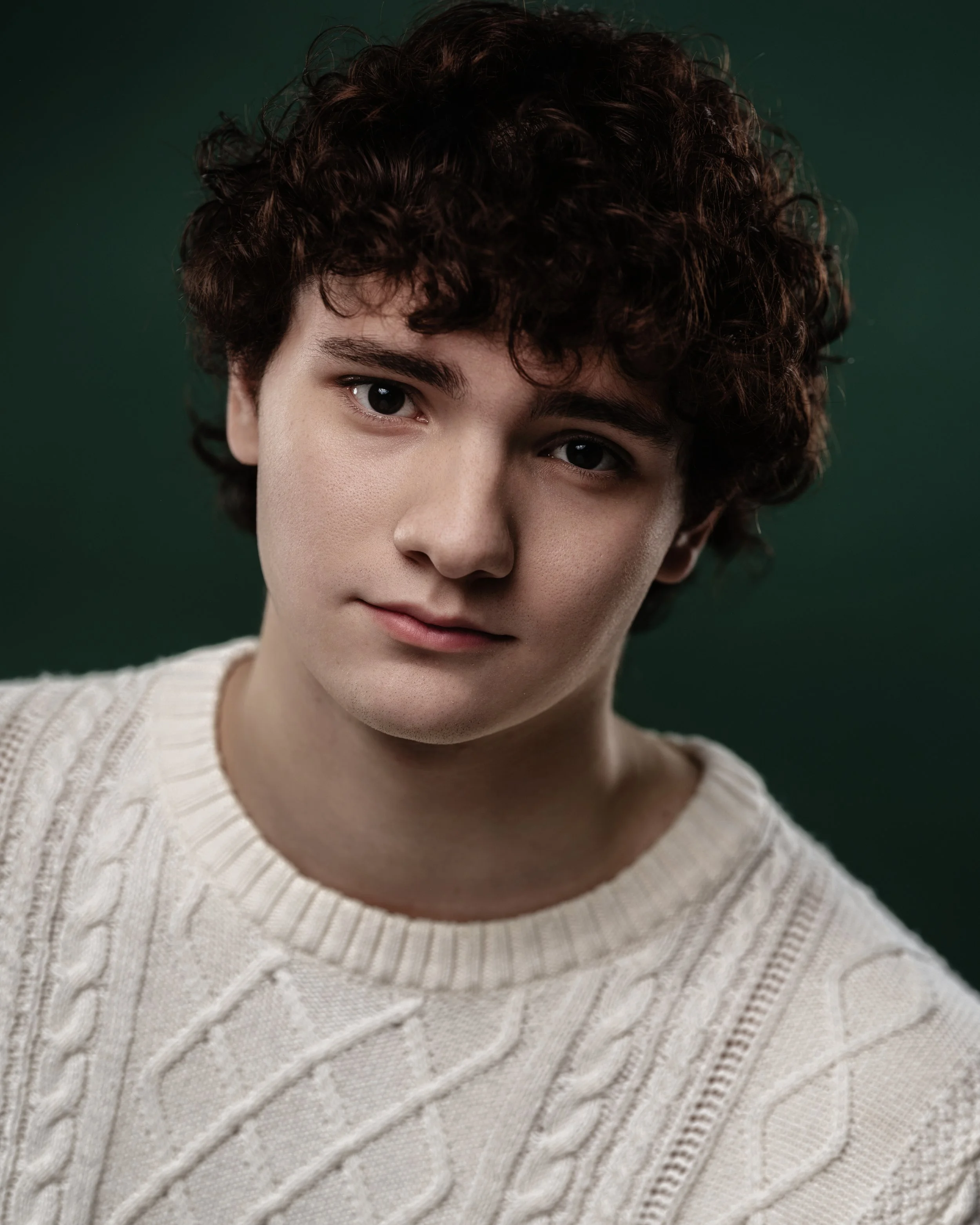 Young person with curly brown hair wearing a cream-colored knitted sweater against a dark green background.