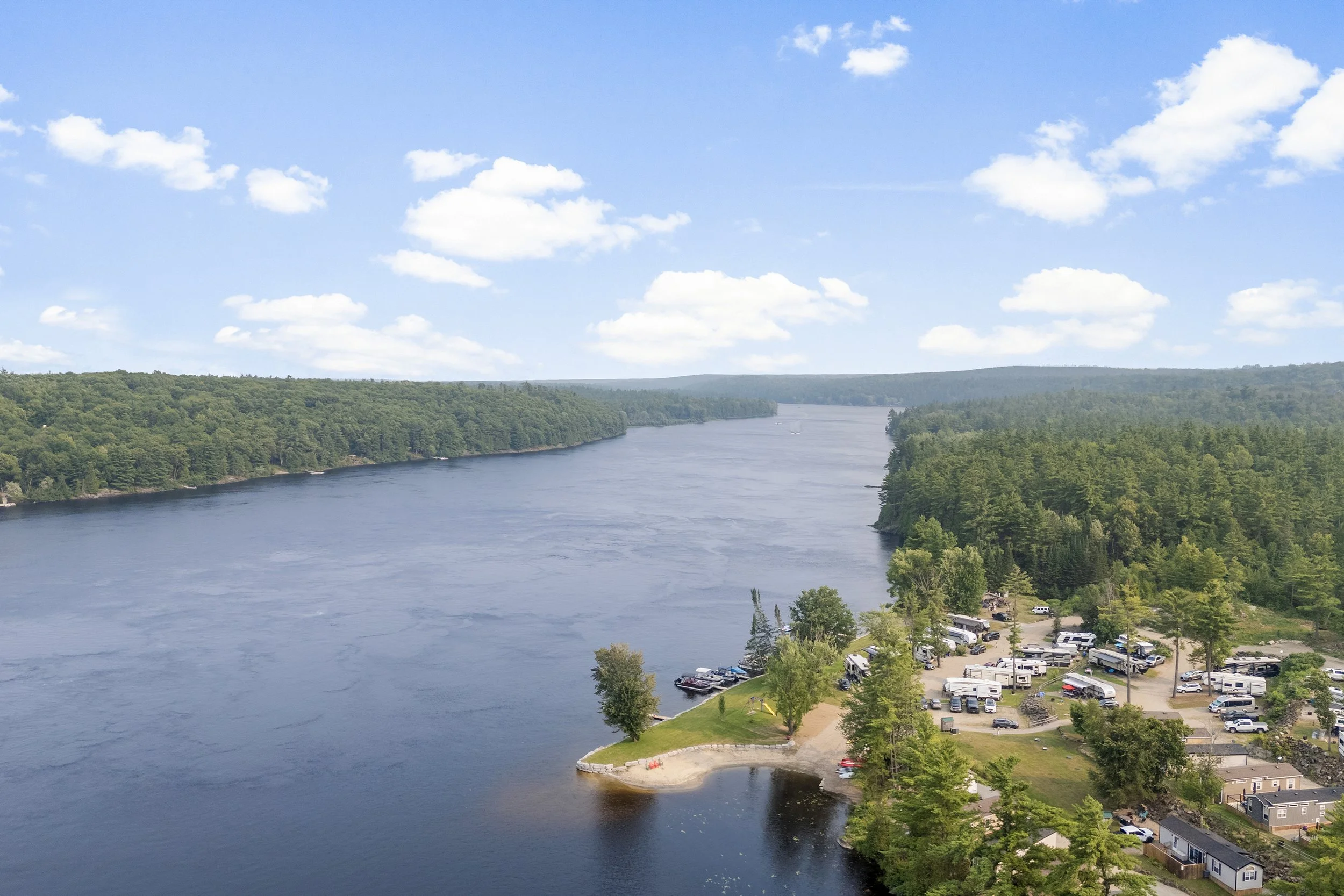 A river flowing through a wooded landscape under a partly cloudy sky, with a campground and RVs along the shore.
