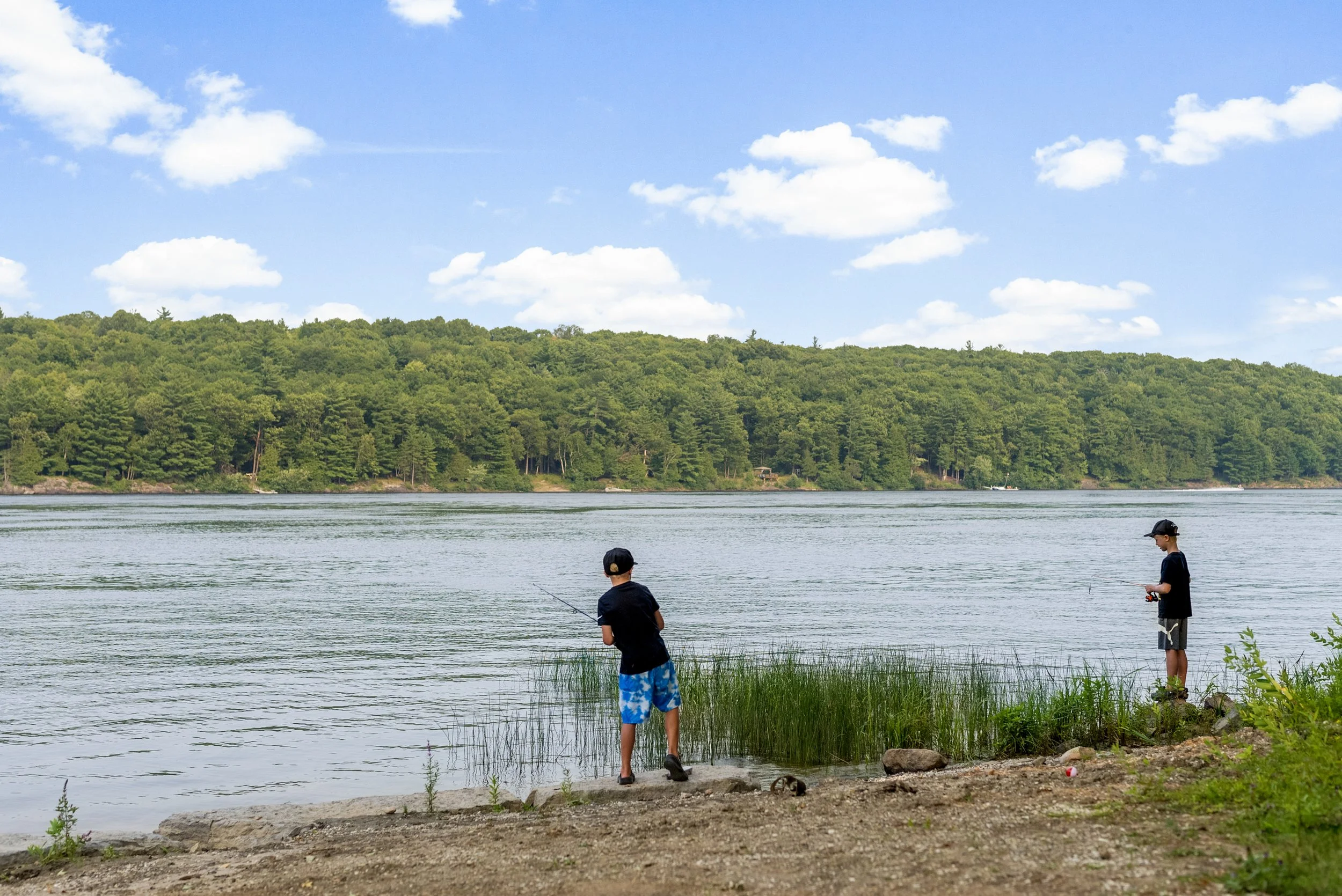 Two children fishing by a lake on a grassy shoreline with green trees and a hill in the background on a partly cloudy day.