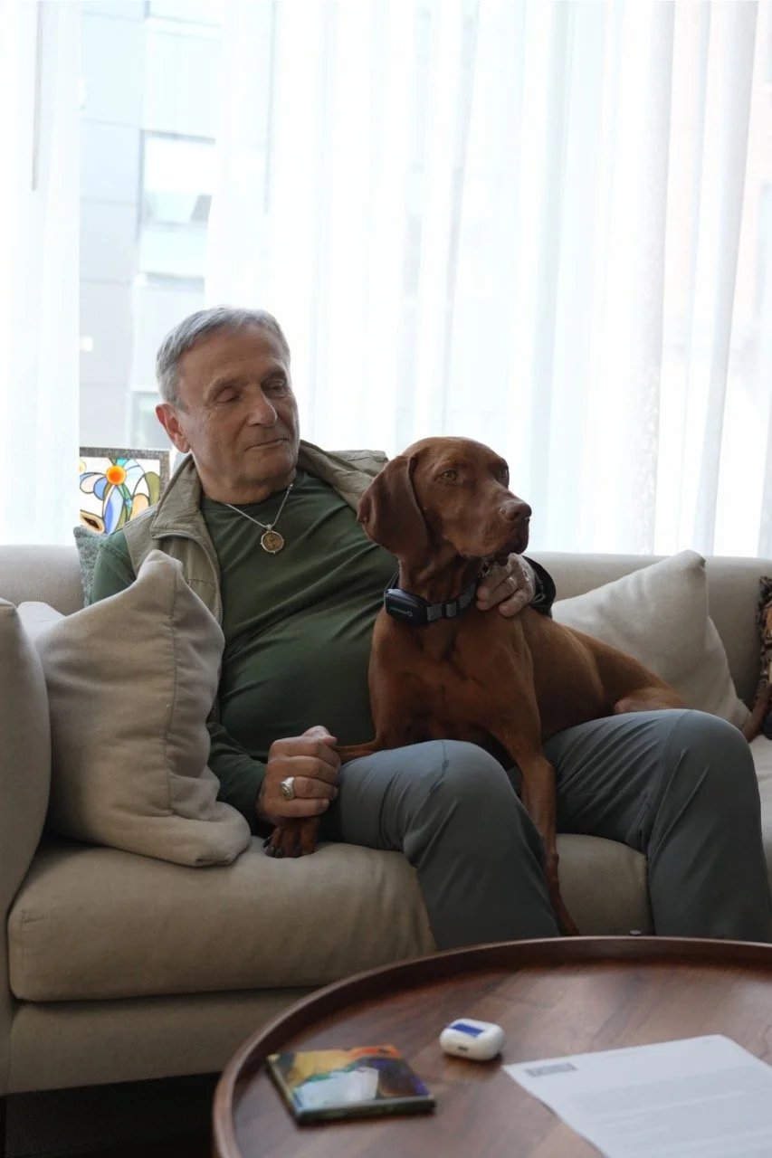 A man sitting on a beige sofa with a brown dog sitting on his lap. The man is wearing a green shirt, a beige vest, and gray pants, with his eyes closed and a pleasant expression. Behind them are sheer white curtains and a stained glass window, with a round wooden table in front of them holding a book, a paper, and a small electronic device.