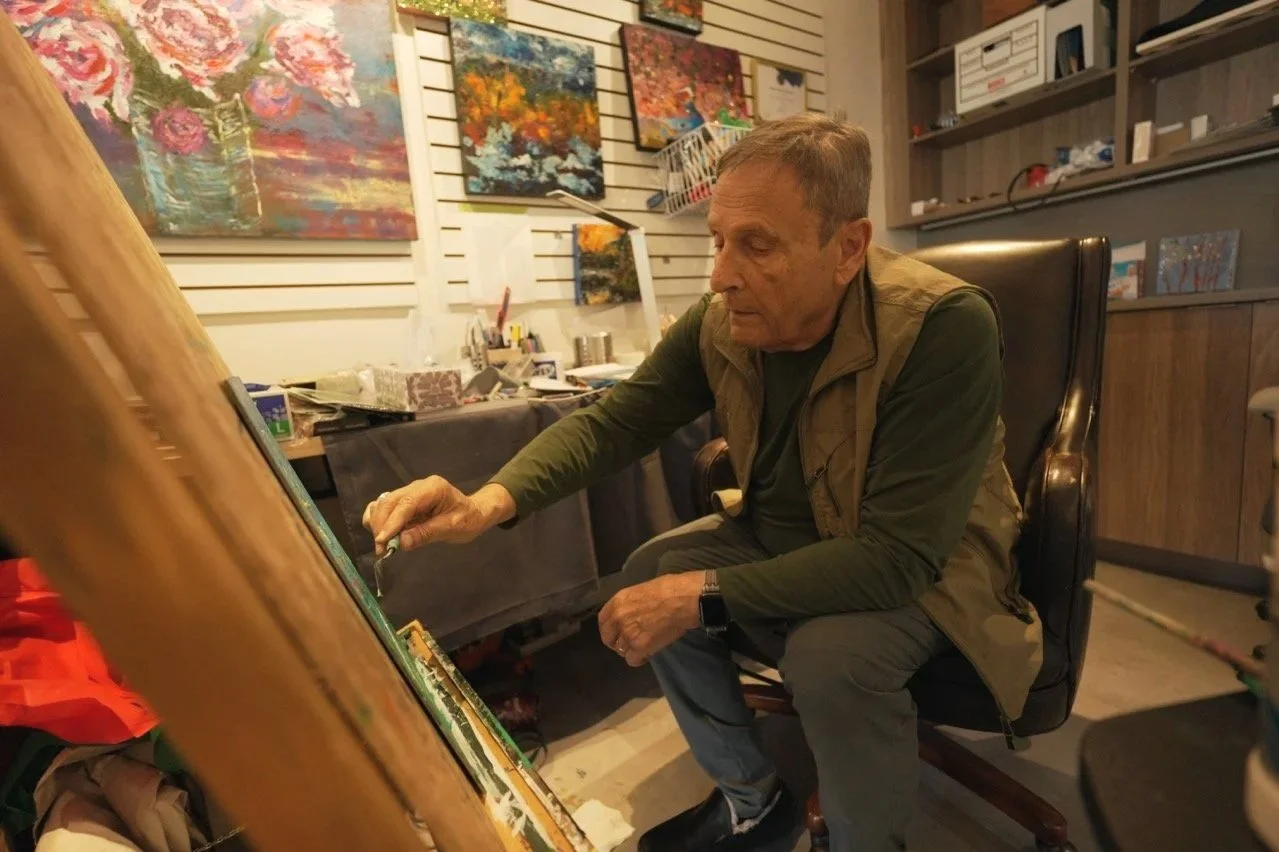 Oscar in a green shirt and brown vest sits on a black chair, painting on a large canvas in an art studio with colourful paintings and art supplies in the background.