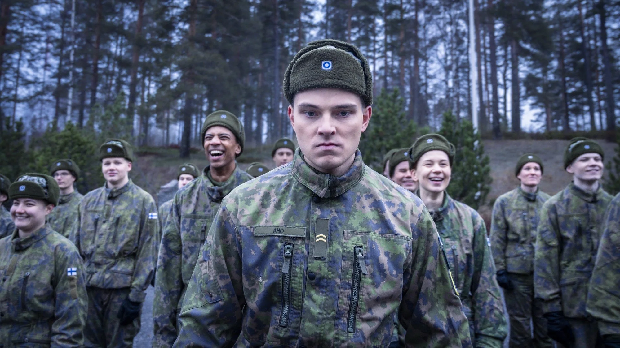 Kuvassa kohtaus Tupa 13-TV-sarjasta. Uudet alokkaat seisovat rivissä ja nauravat. Alikersanttia ei hymyilytä..