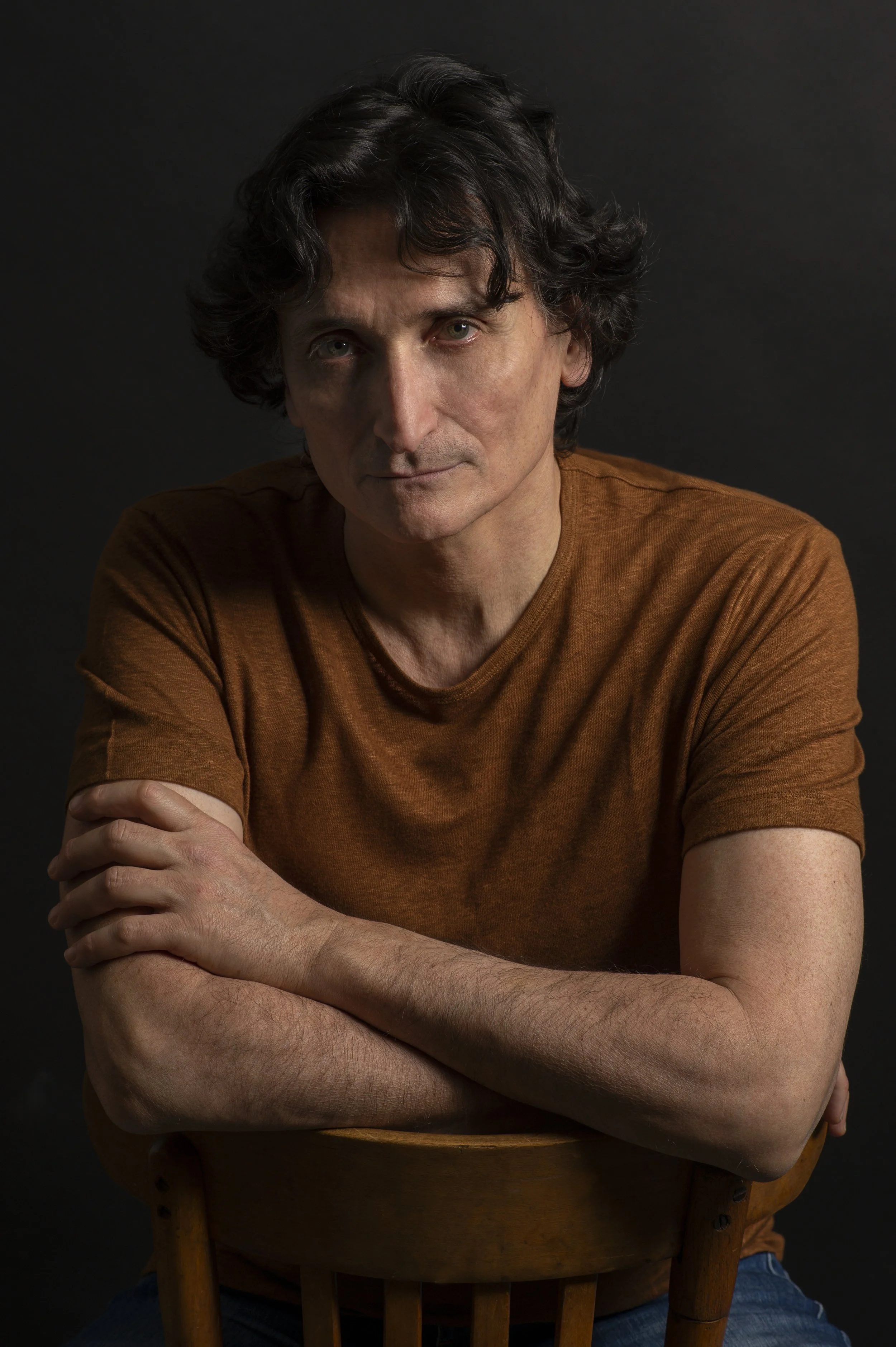 Portrait d'un homme avec des cheveux bouclés, portant un t-shirt marron, sur un fond noir, assis sur une chaise en bois.