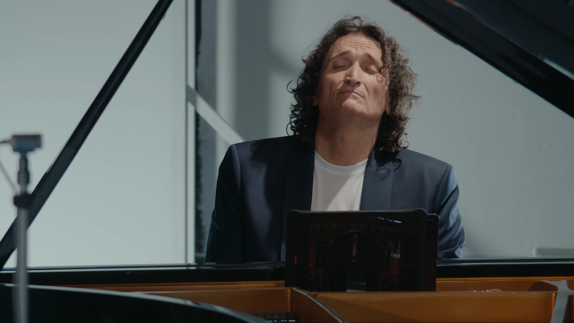 Un homme aux cheveux bouclés joue du piano, portant un veston noir et une chemise blanche, avec une expression de concentration ou de passion.