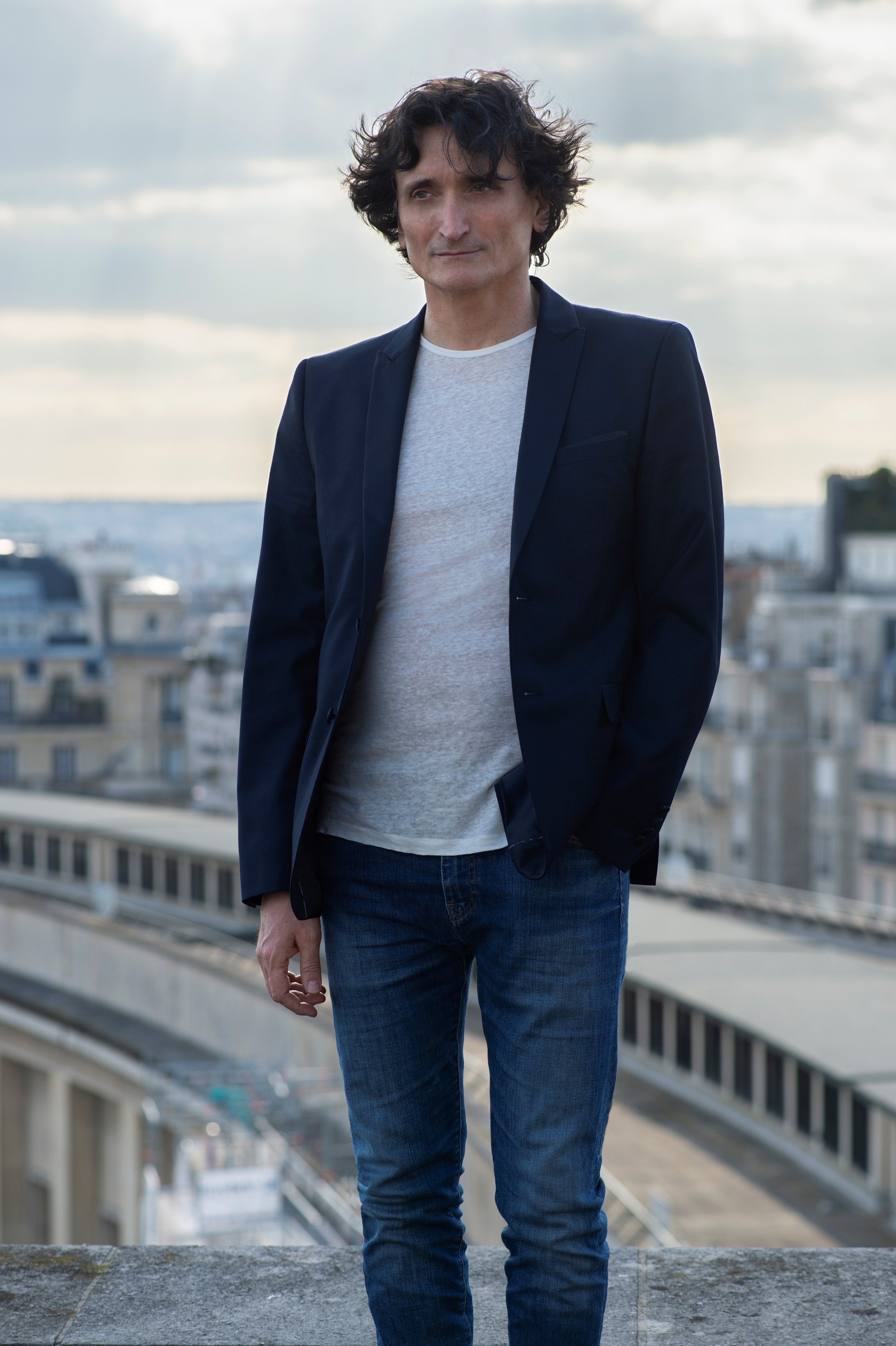 Un homme avec des cheveux noirs bouclés, portant une veste bleue marine, un t-shirt gris, et un jean, se tient sur un toit avec une vue urbaine en arrière-plan, sous un ciel nuageux.