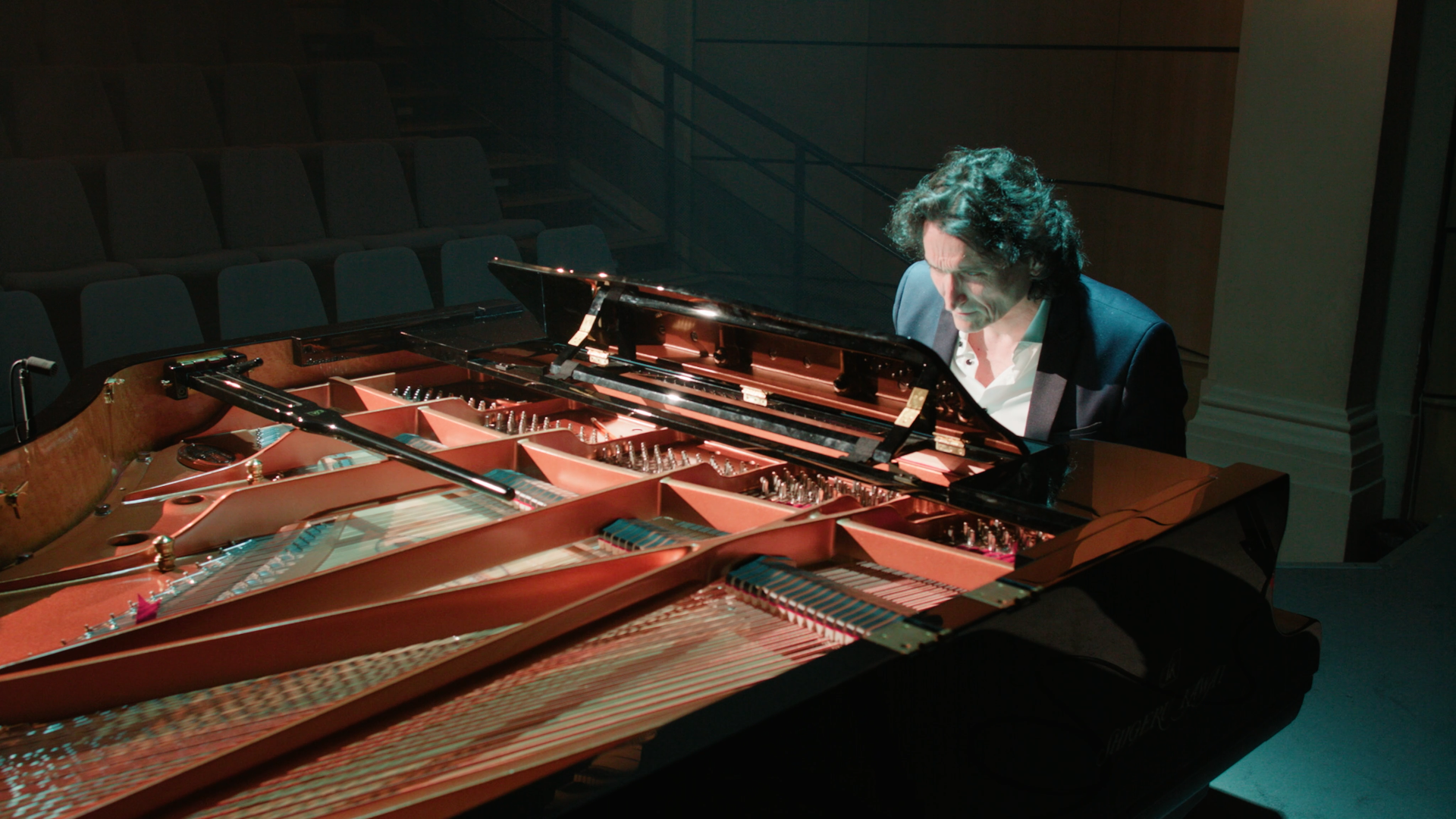 Un homme joue du piano à queue dans une salle de concert vide, éclairé par une lumière tamisée.