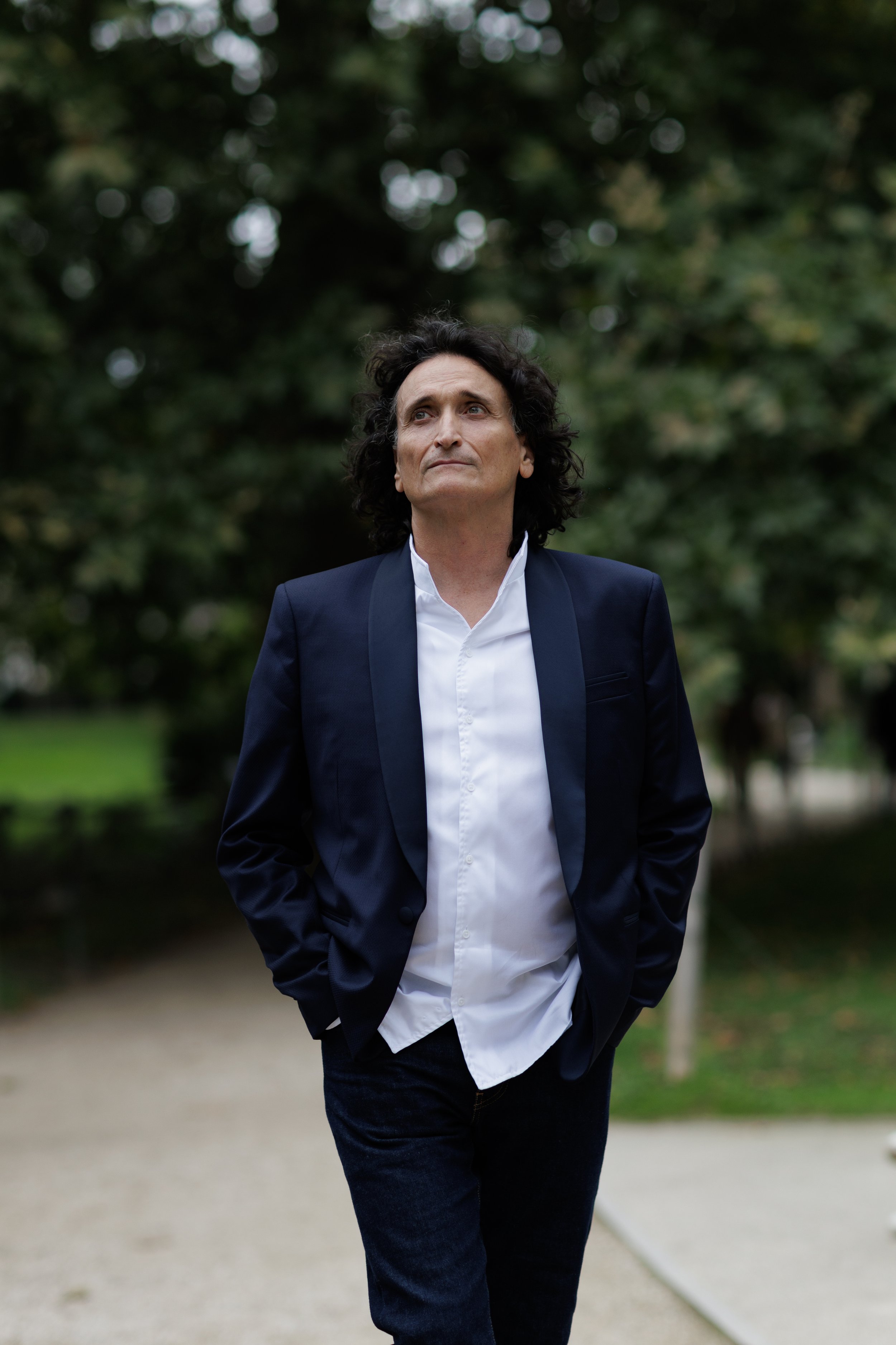 Un homme en costume noir, chemise blanche, marche dans un parc, mains dans les poches, regard vers le haut, avec des arbres en arrière-plan.