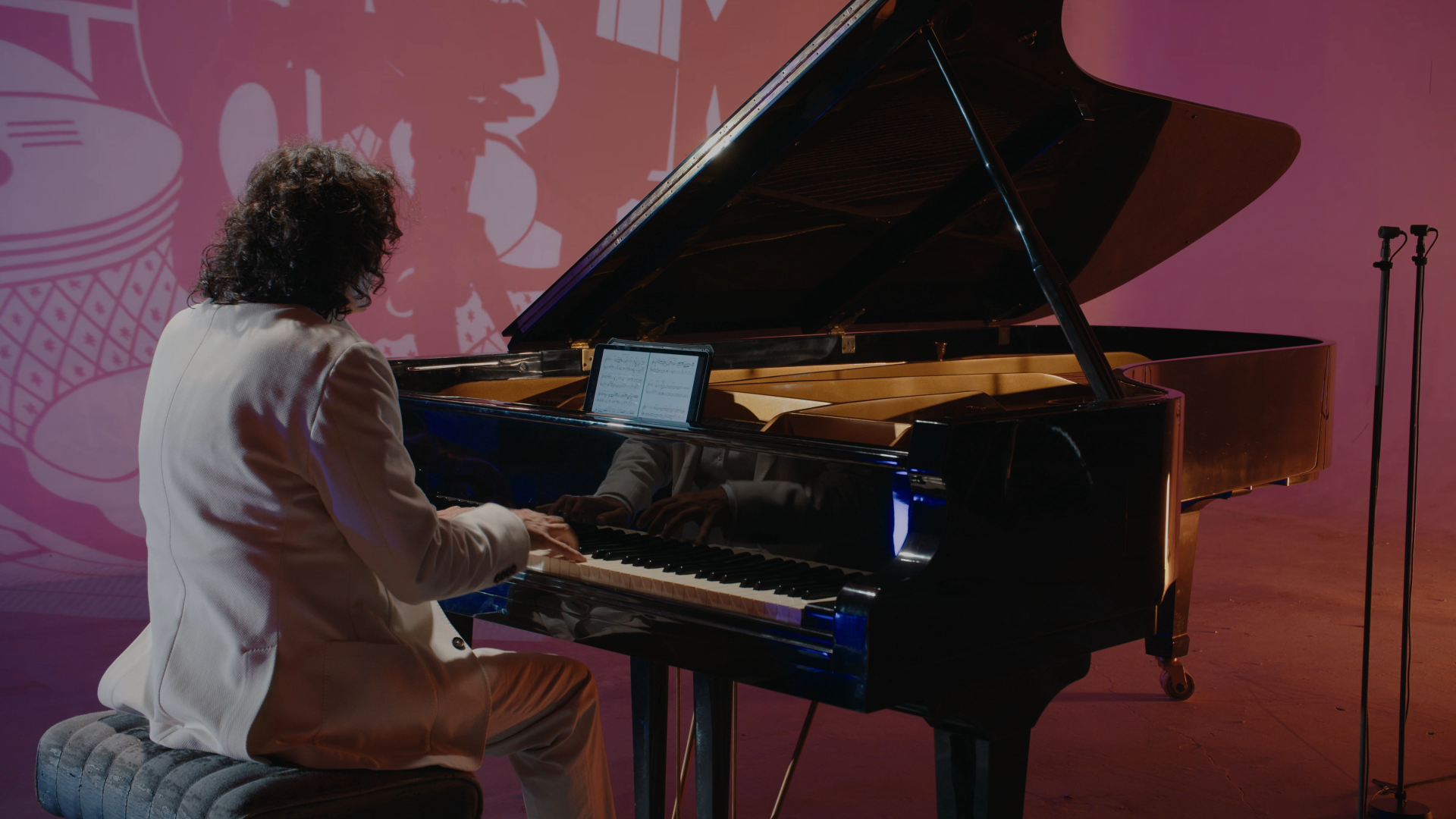 Personne jouant du piano à queue noir, en robe blanche, avec une tablette affichant des partitions à l'écran, dans un décor coloré avec un fond rose et des micros sur pied.