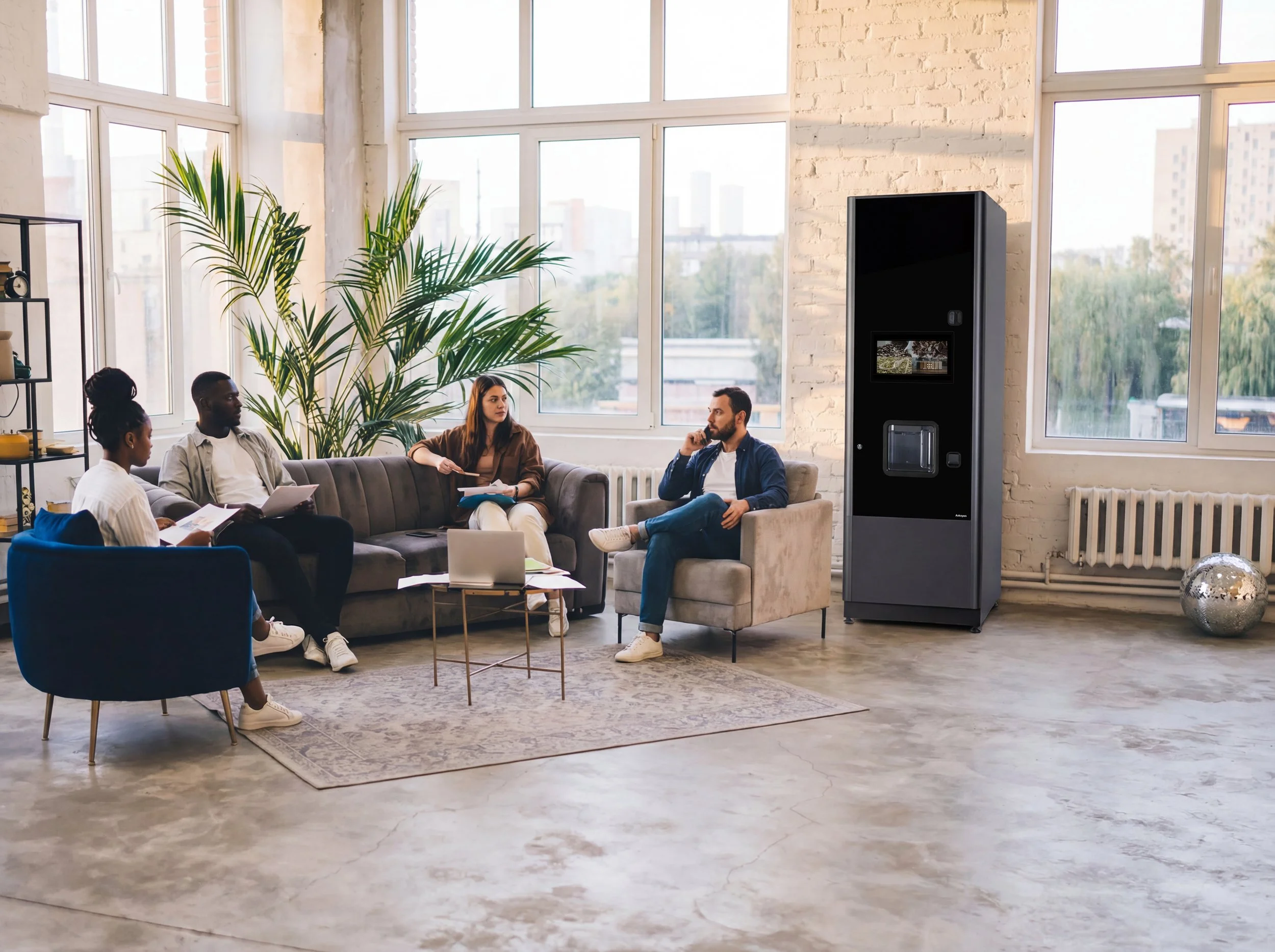 Vier Personen sitzen in einem modernen, hellen Raum mit großen Fenstern, vollen Pflanzen und einer Wand aus weißen Ziegelsteinen. Sie führen eine lockere Diskussion geführt werden, mit einem Laptop, Notizblättern und einem Getränkeautomat im Hintergrund.