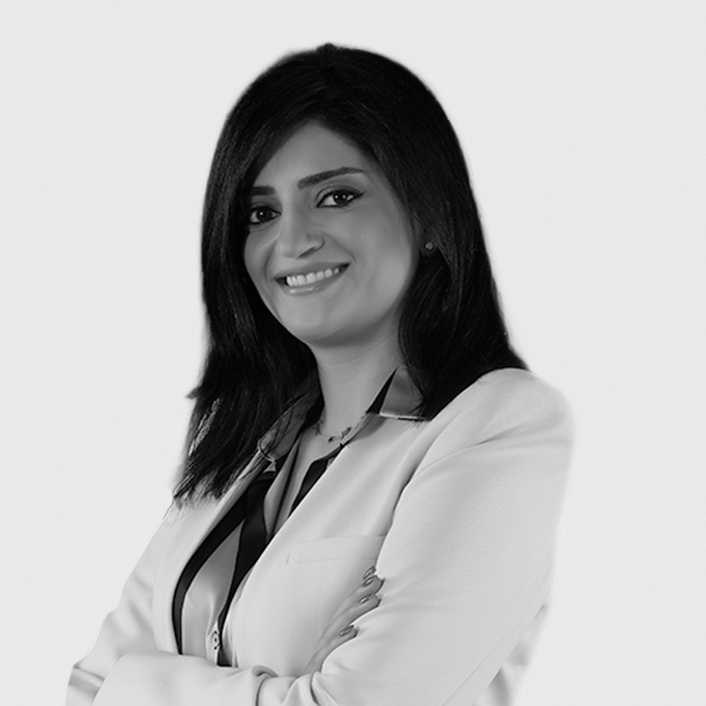 Black and white portrait of a woman with dark hair, wearing a black blazer and a patterned top, standing against a plain background.