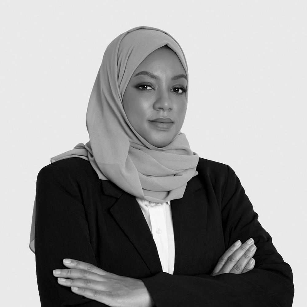 Black and white portrait of a woman with shoulder-length dark hair, wearing a dark blazer, standing with her arms crossed against a plain light background.