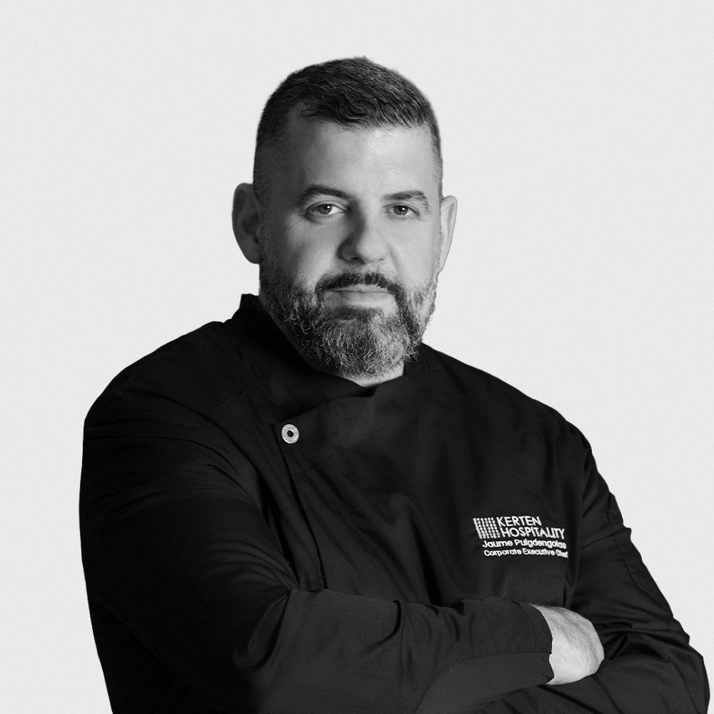 Black and white portrait of a man with a beard, crossing his arms, smiling slightly, wearing a zip-up sweater over a T-shirt, with a neutral background.