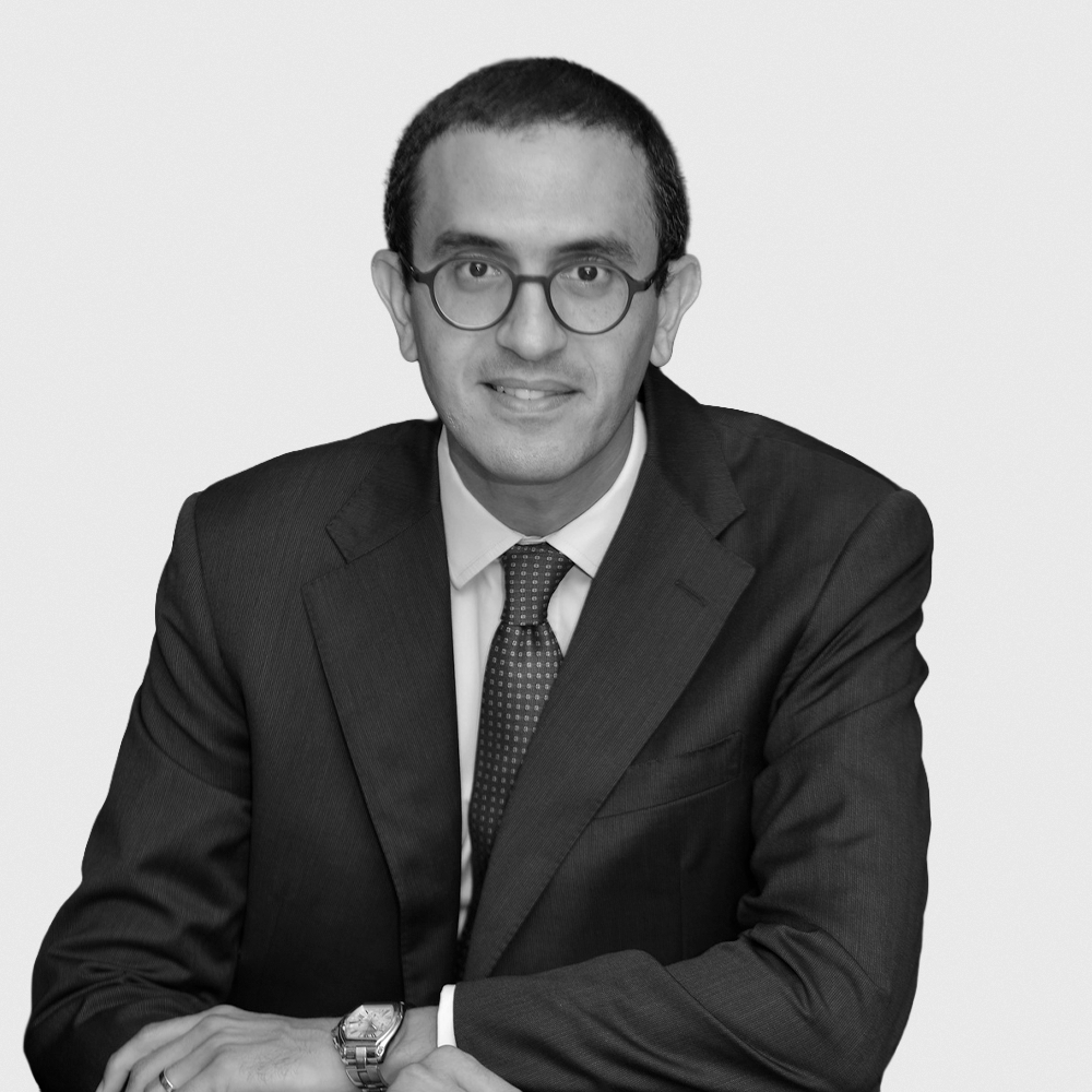 A black-and-white portrait of a man wearing glasses, a suit, and a tie, looking directly at the camera with a slight smile.