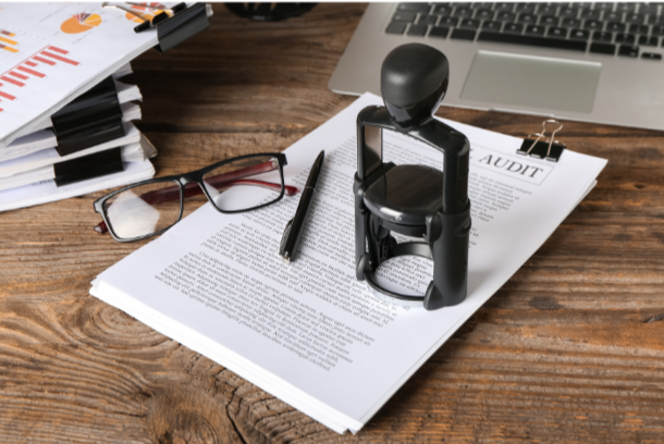 Audit document on a wooden desk with stamp, glasses, pen, and laptop.