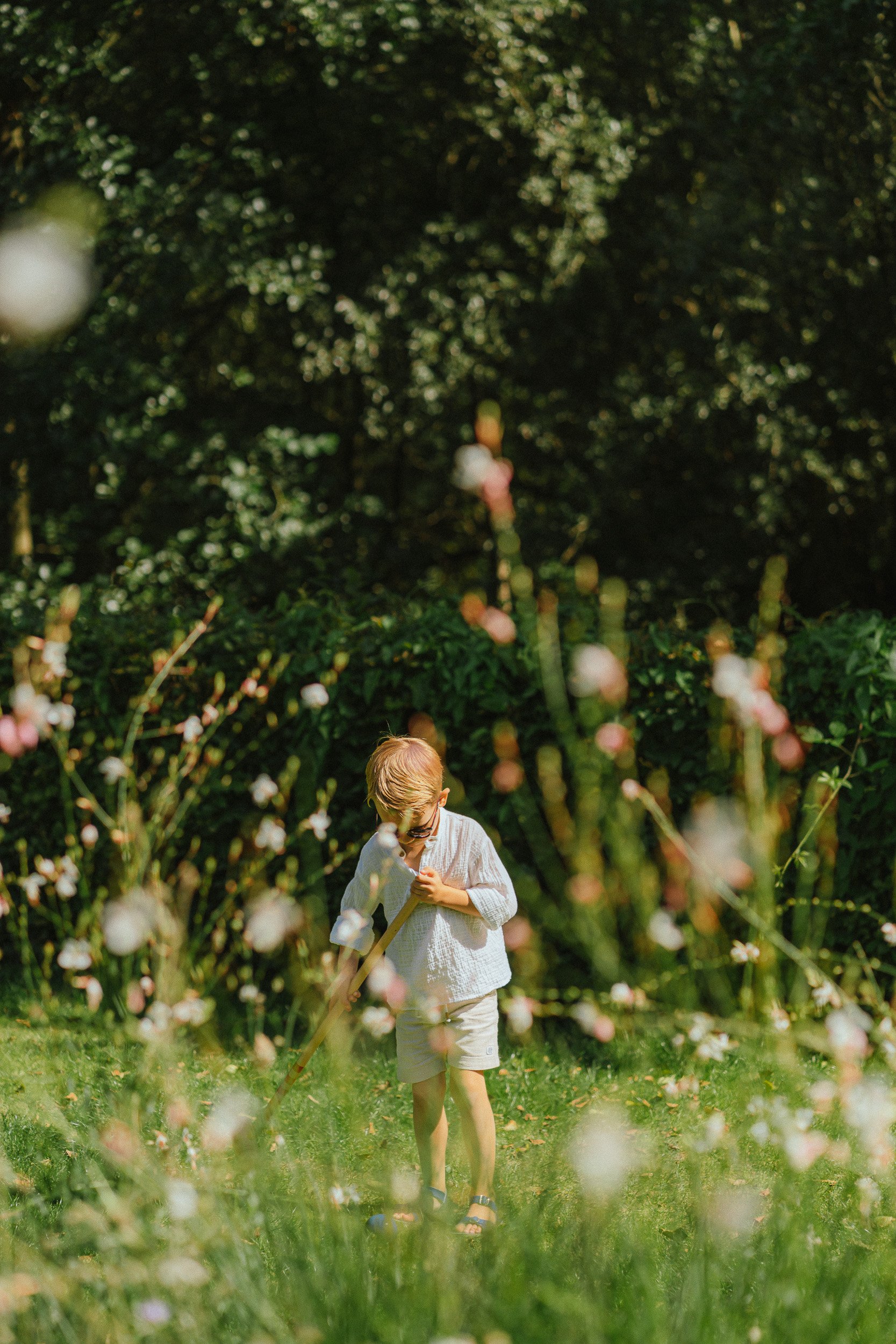 Een kind, gekleed in een wit overhemd en korte broek, dat met een hark in een bloemenveld werkt, omringd door bloemen en groen.