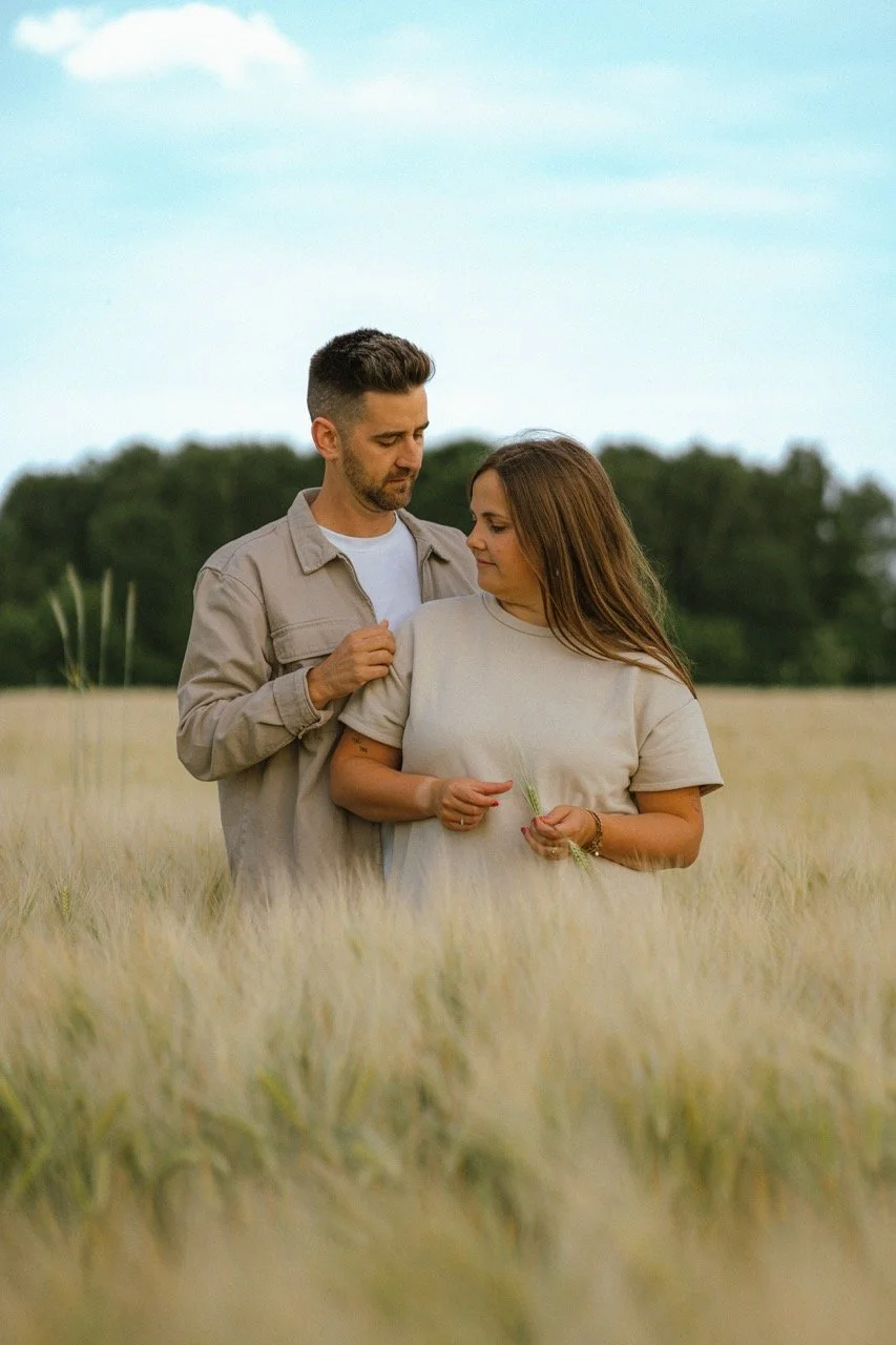 Een man en een vrouw staan dicht bij elkaar in een weiland, de man vasthoudt de schouder van de vrouw en ze staan omringd door gras, met een groene boomgaard en een blauwe lucht op de achtergrond.