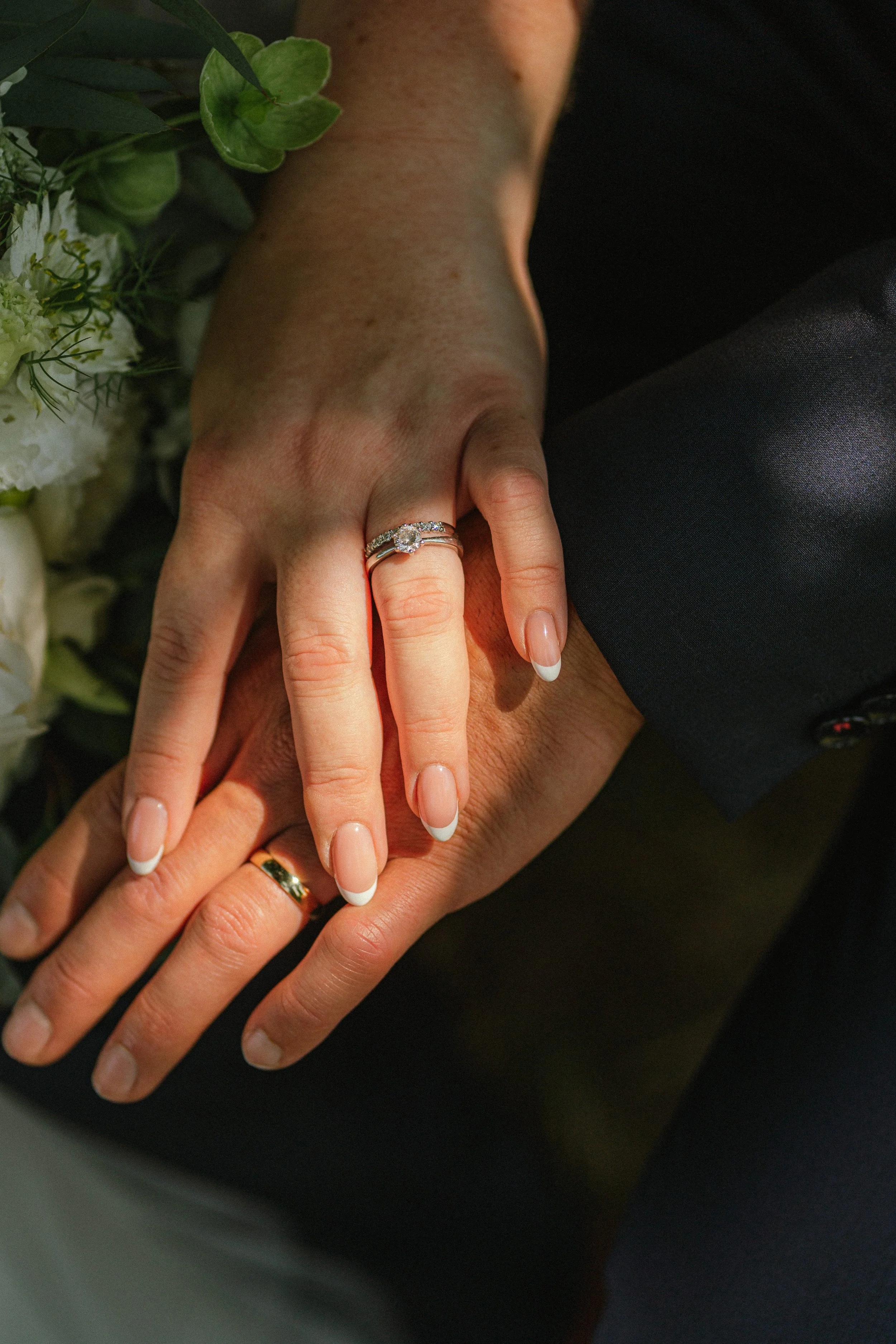 Close-up van twee handen met elkaar vastgehouden, een ring met een grote diamant op de ringvinger, en een bloemenarrangement op de achtergrond.