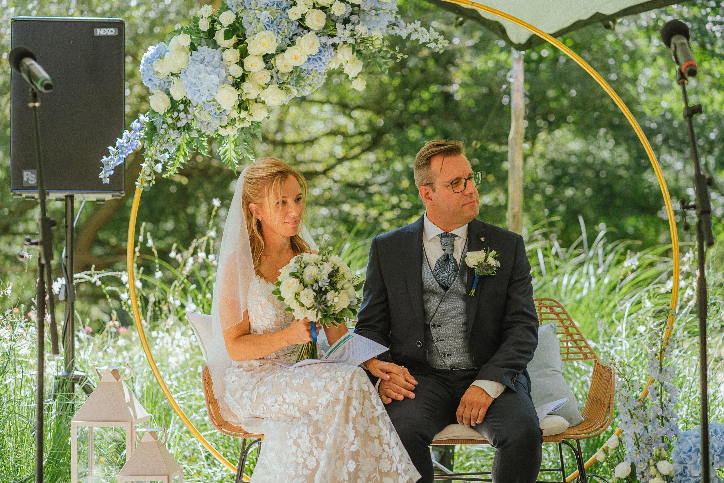 Een bruid en bruidegom tijdens hun huwelijk, zittend onder een bloemrijke archel bij een buitenceremonie.