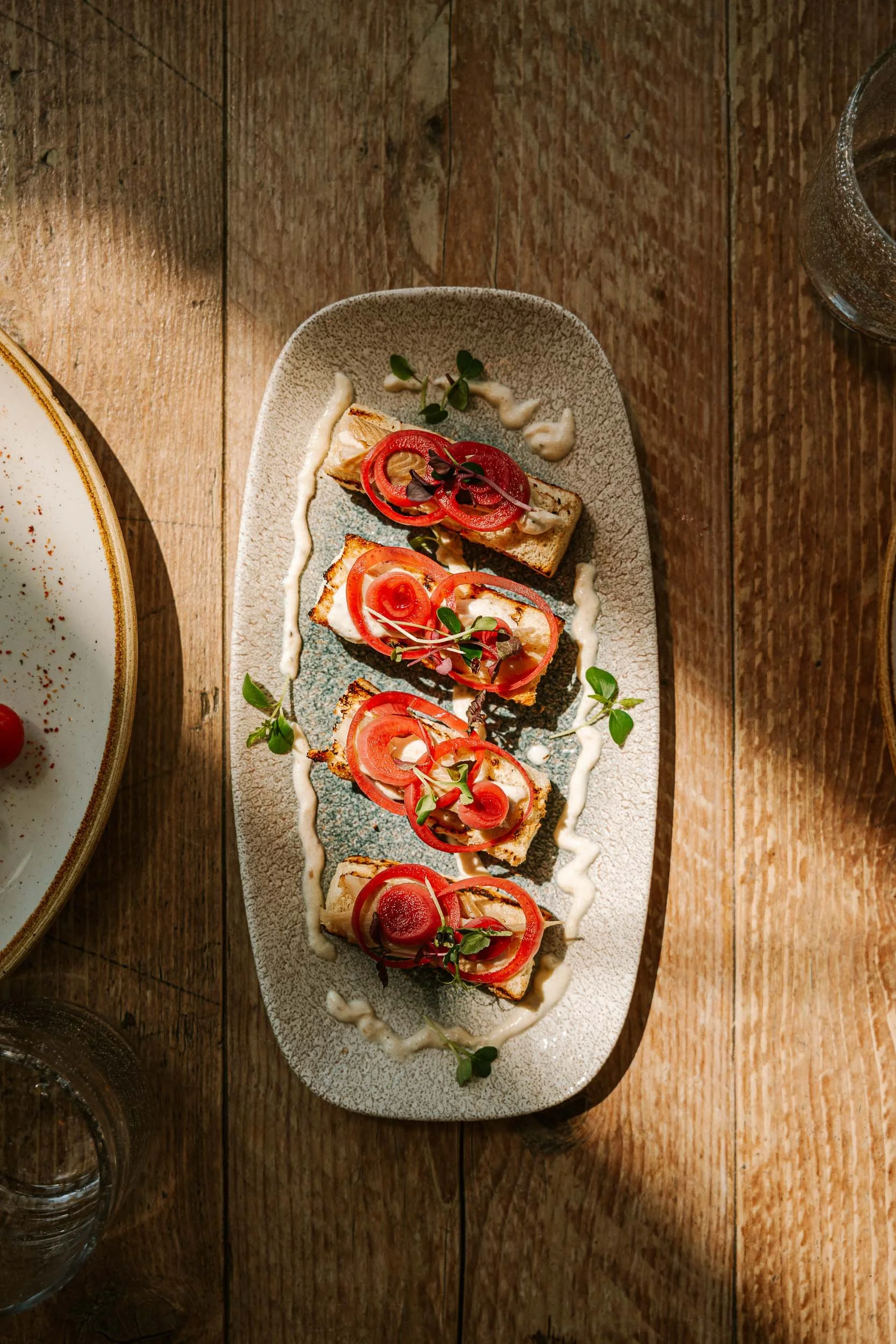 Geregen toast met tomaat, radijs, en microgreens op een langwerpig bord, geserveerd op een houten tafel.